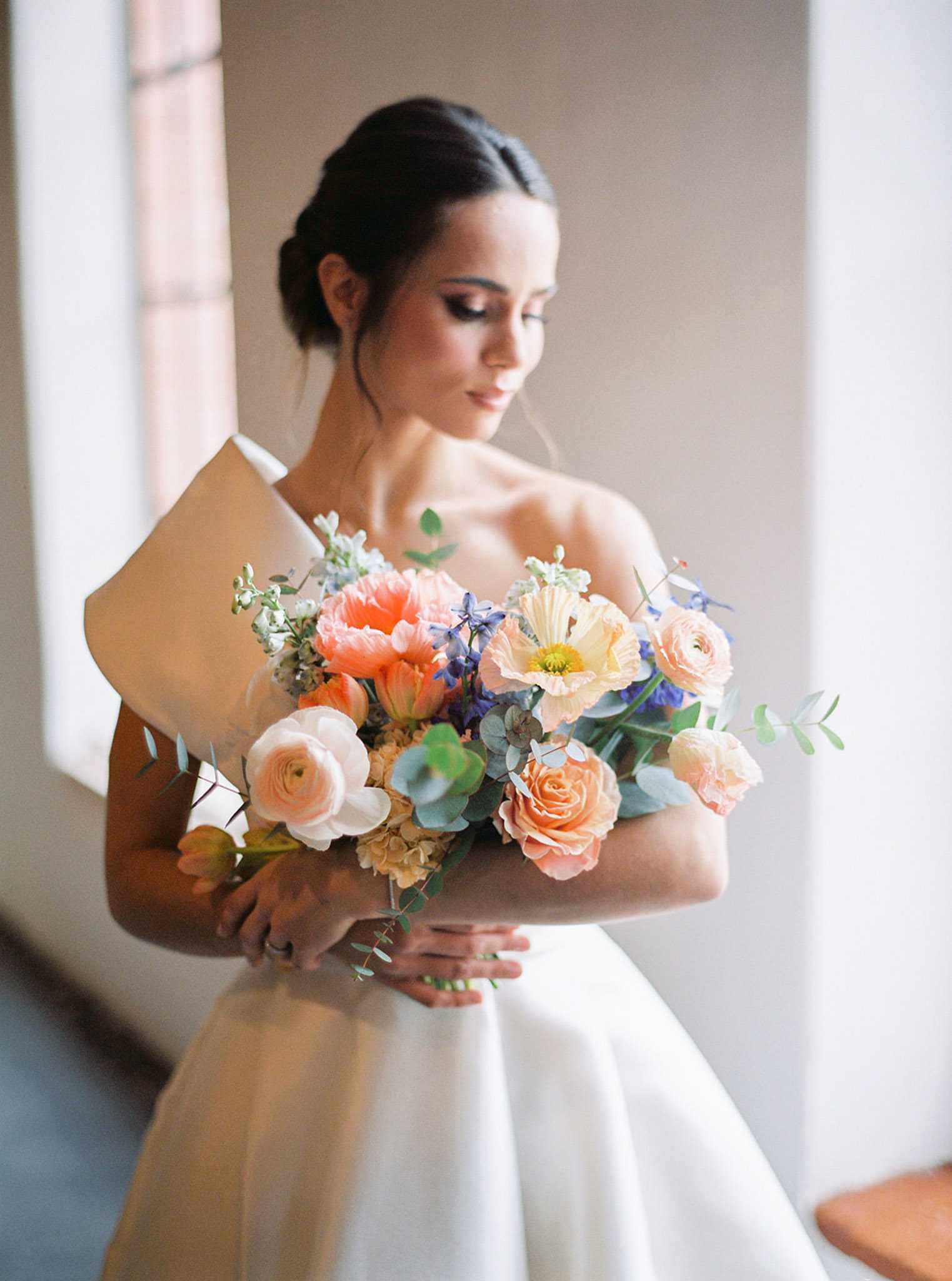 A bridal portrait taken indoors near a window, showing a single bride holding a large, loosely arranged bouquet wrapped in kraft paper. The bride wears a strapless or off-shoulder white gown with a structured neckline and has her dark hair pulled back in a low updo; she looks downward with eyes slightly closed. The bouquet features coral and peach poppies, peach and blush ranunculus, peach garden roses, orange tulips, periwinkle-blue delphinium, cream hydrangea, white stock flowers, and eucalyptus foliage in a garden-style arrangement with a warm coral, peach, and blue color palette. The composition is a medium portrait shot with soft natural window light and a neutral, blurred interior background.