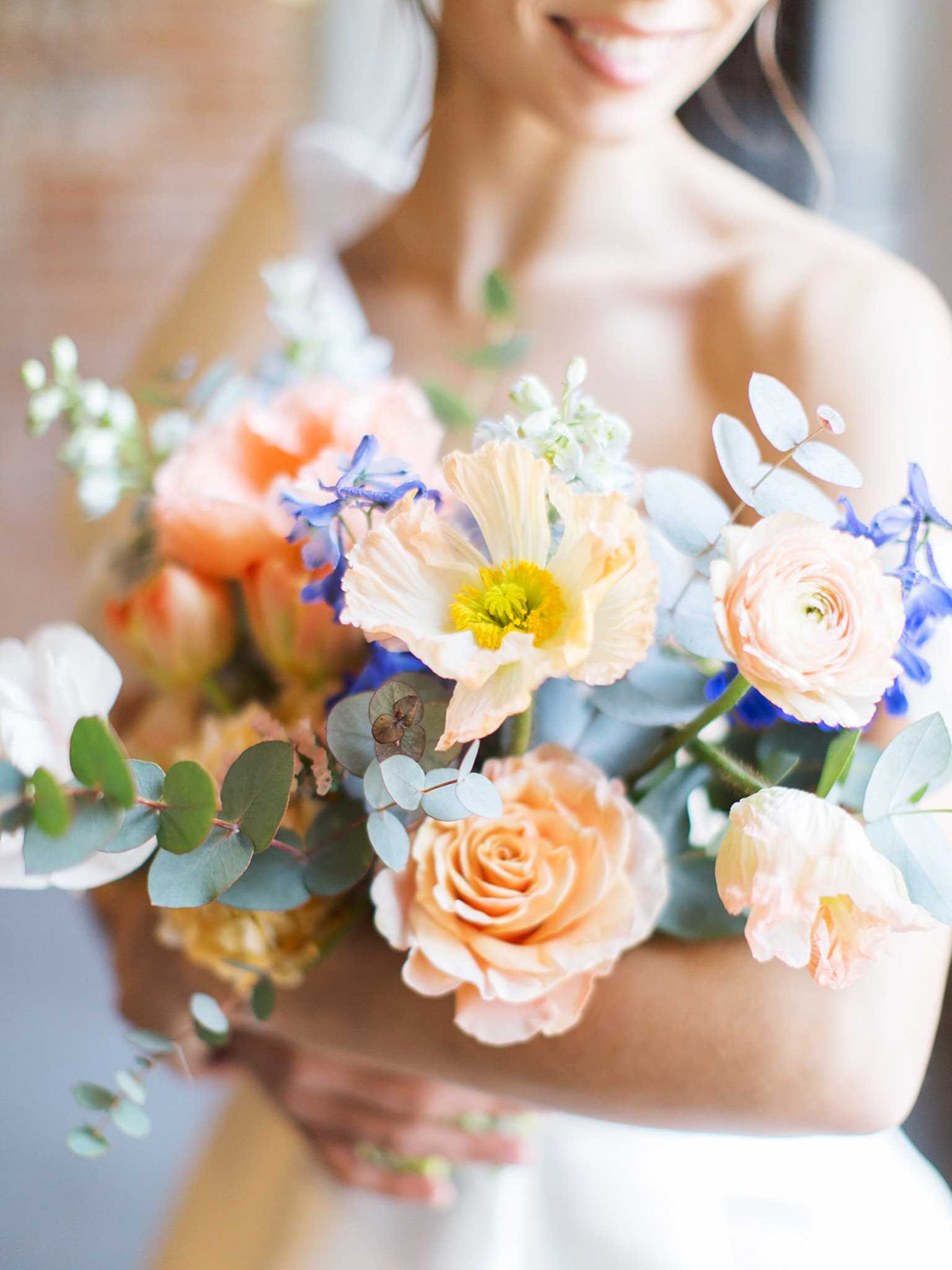 A close-up detail shot of a bride holding her bridal bouquet, with the bride's face softly out of focus in the background. The bride is wearing a white strapless gown and has dark hair. The bouquet features a mix of peach garden roses, peach ranunculus, a cream poppy with a yellow center, cobalt blue delphinium, soft peach tulips, white stock flowers, and trails of silver-dollar eucalyptus, creating a warm peach-and-cobalt blue color palette with greenery accents. The composition is a tight portrait-style detail shot with a shallow depth of field that draws focus entirely to the bouquet's floral arrangement.