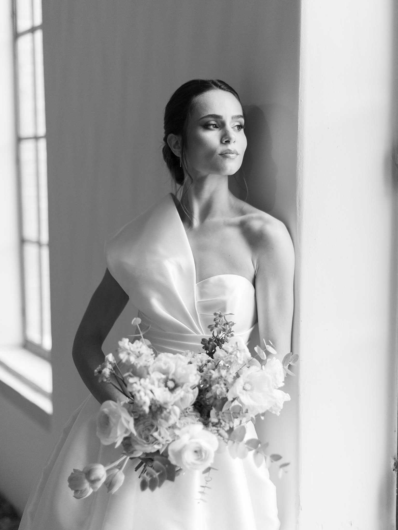 This is a black-and-white bridal portrait shot indoors, with the bride standing against a wall beside a large multi-pane window that casts soft directional light across her face and dress. The bride wears a structured strapless gown with a dramatic folded or draped detail at the bodice, styled in a sleek, modern silhouette. Her dark hair is pulled back into a low updo, and she gazes off to one side with a composed expression. She holds a loose, garden-style bouquet featuring what appear to be anemones, ranunculus, tulips, and eucalyptus, with varied light and dark tones suggesting a mix of pale and deeper-toned blooms. The composition is a close-up portrait with strong contrast between the bright window light and the shadowed background, emphasizing the clean lines of the dress and the texture of the florals.