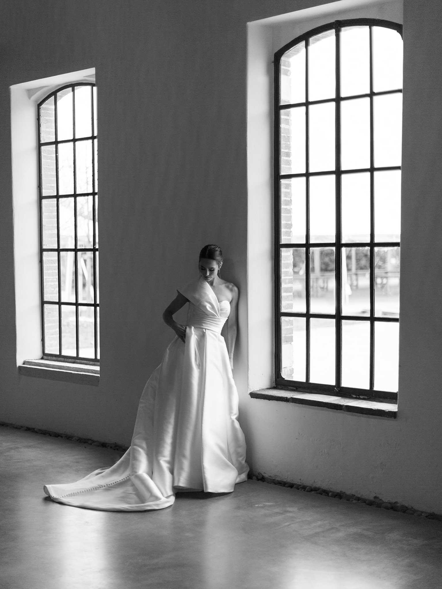 This black-and-white bridal portrait shows a bride standing alone indoors between two tall arched steel-framed windows that flood the space with bright natural light, creating strong contrast against the pale plastered walls and polished concrete floor. The bride wears a structured, minimalist satin ball gown with a sweetheart neckline, a single asymmetric off-shoulder strap, and a long cathedral-length train spread across the floor behind her. Her hair is pulled back into an updo and she gazes downward with one hand resting on her hip. The composition is a full-length portrait shot taken from a slight distance, emphasizing the architectural industrial-style interior and the clean lines of the modern gown.