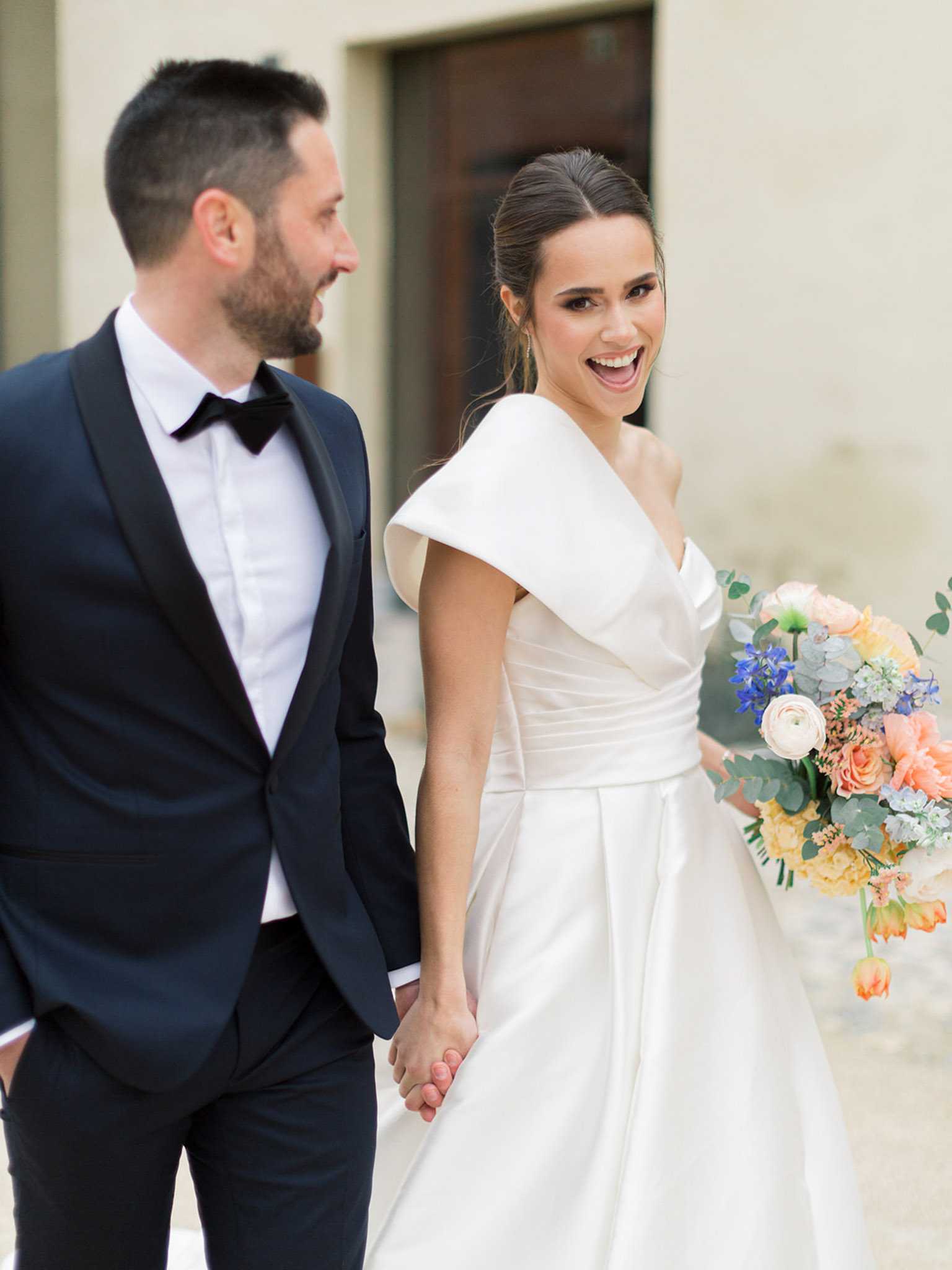 A couple portrait taken outdoors against a light stone building facade, with the bride and groom holding hands and walking. The groom wears a navy tuxedo with black satin lapels, a white dress shirt, and a black bow tie. The bride wears a white structured satin ball gown with a distinctive wide off-the-shoulder portrait collar and a fitted sash waist, her dark hair pulled back in a half-updo; she is laughing and looking toward the camera. She carries a colorful bouquet featuring peach ranunculus, coral roses, orange tulips, yellow carnations, cobalt blue delphinium, pale blue flowers, and eucalyptus foliage. The overall styling is modern and formal. Medium portrait shot, slightly shallow depth of field.