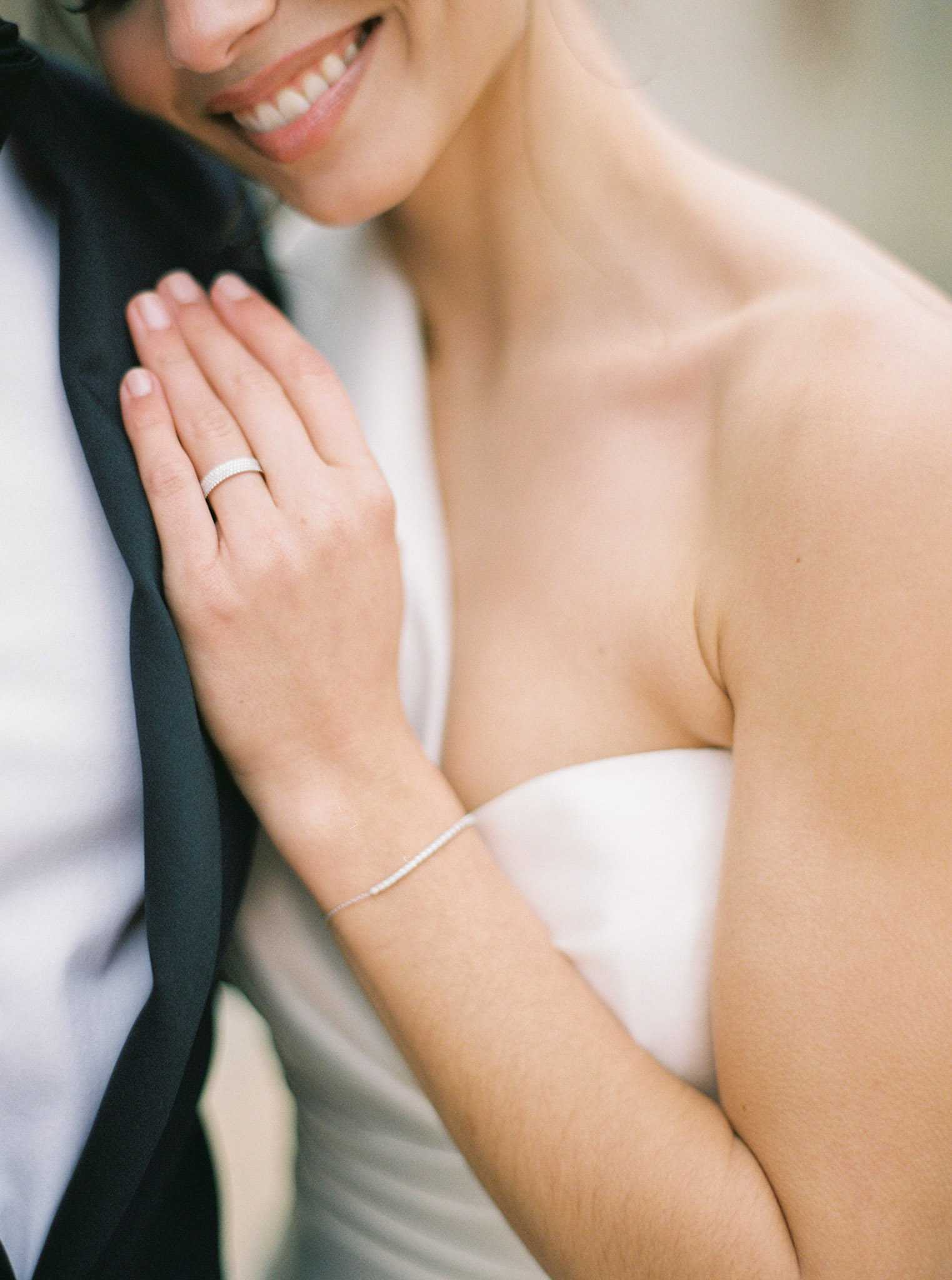 A close-up detail portrait of a bride leaning against a groom dressed in a black tuxedo with a white dress shirt. The bride wears a strapless white gown with a smooth satin finish, a delicate diamond or crystal tennis bracelet on her wrist, and a pavé diamond wedding band on her ring finger. Her nails are finished with a natural nude polish. The composition is tightly cropped, showing only the lower half of the bride's smiling face, her hand resting flat against the groom's lapel, and her bare shoulder and décolletage. The styling is clean and modern with a neutral color palette of white and black. Shot with a shallow depth of field against a softly blurred neutral background.