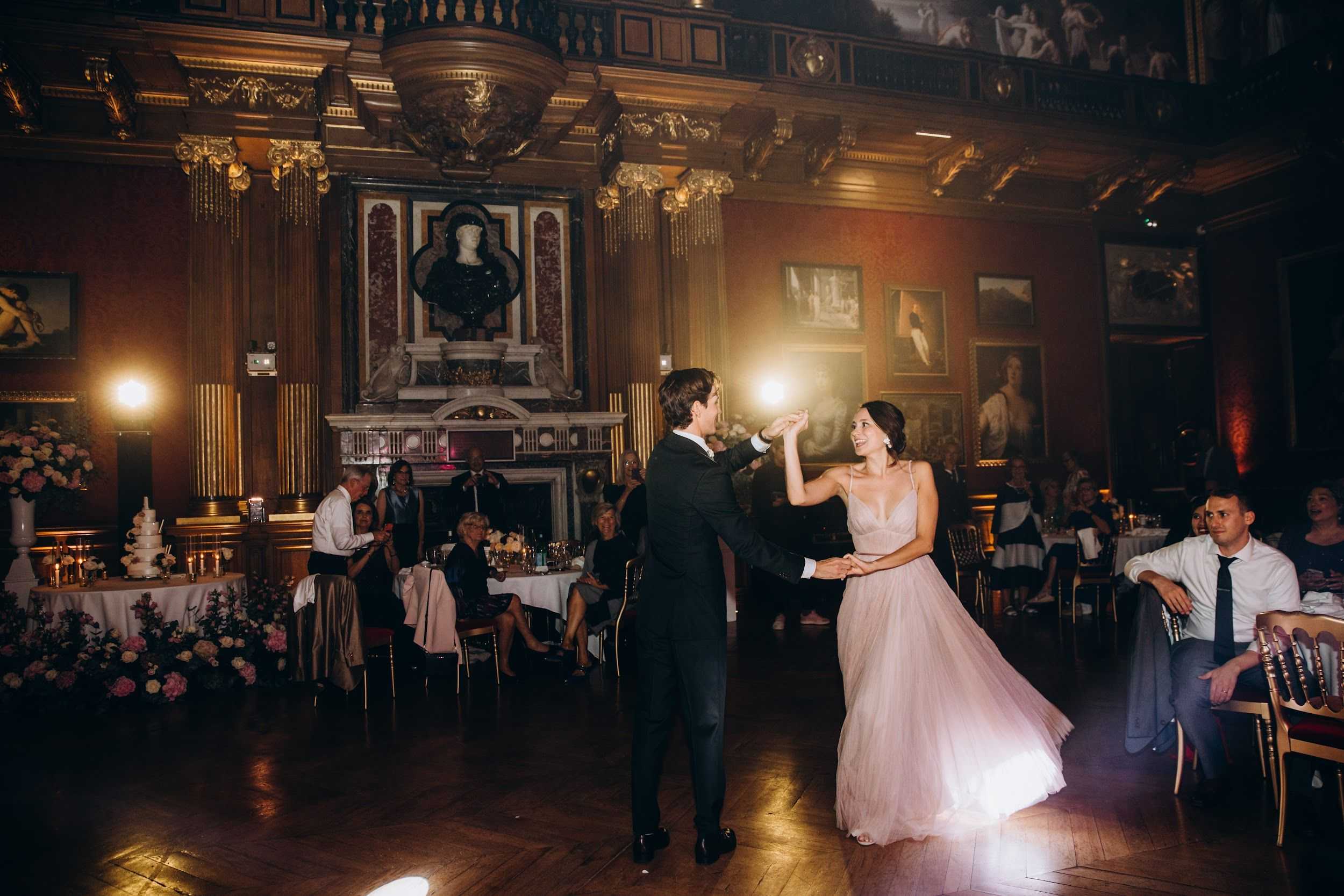 The couple shares their first dance in the center of a grand indoor ballroom featuring dark wood paneling, gilded Corinthian columns, oil paintings on the walls, an ornate marble fireplace with a classical bust above it, and painted ceiling medallions — consistent with a historic European palace or stately home. The groom wears a dark navy suit while the bride wears a blush pink tulle ball gown with spaghetti straps, her hair styled in an updo. He is spinning her as she smiles, lit by warm amber uplighting and a bright spotlight. Approximately 20–30 seated guests watch from round tables dressed in white linens with gold chiavari chairs; a tiered white wedding cake is visible on the left beside low arrangements of pink, white, and blush florals including roses. The shot is a wide-angle full-length image capturing both the couple and the full room environment. Potential venue feature image.