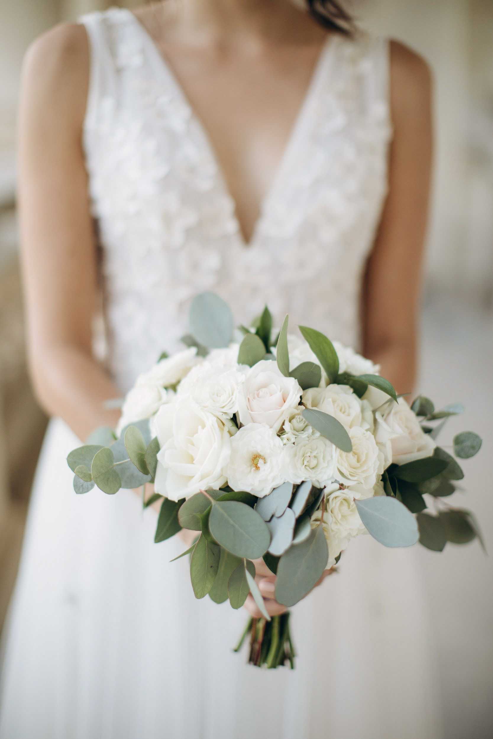 A close-up portrait of a bride holding her bouquet toward the camera, with the bouquet in sharp focus and the bride's torso softly blurred in the background. The bouquet is a rounded arrangement of ivory and blush roses mixed with open garden roses and silver-dollar eucalyptus, tied with a simple stem wrap. The bride wears a white sleeveless gown with a deep V-neckline and three-dimensional floral appliqué detailing on the bodice. The background is soft and neutral, suggesting an indoor setting with diffused natural light.