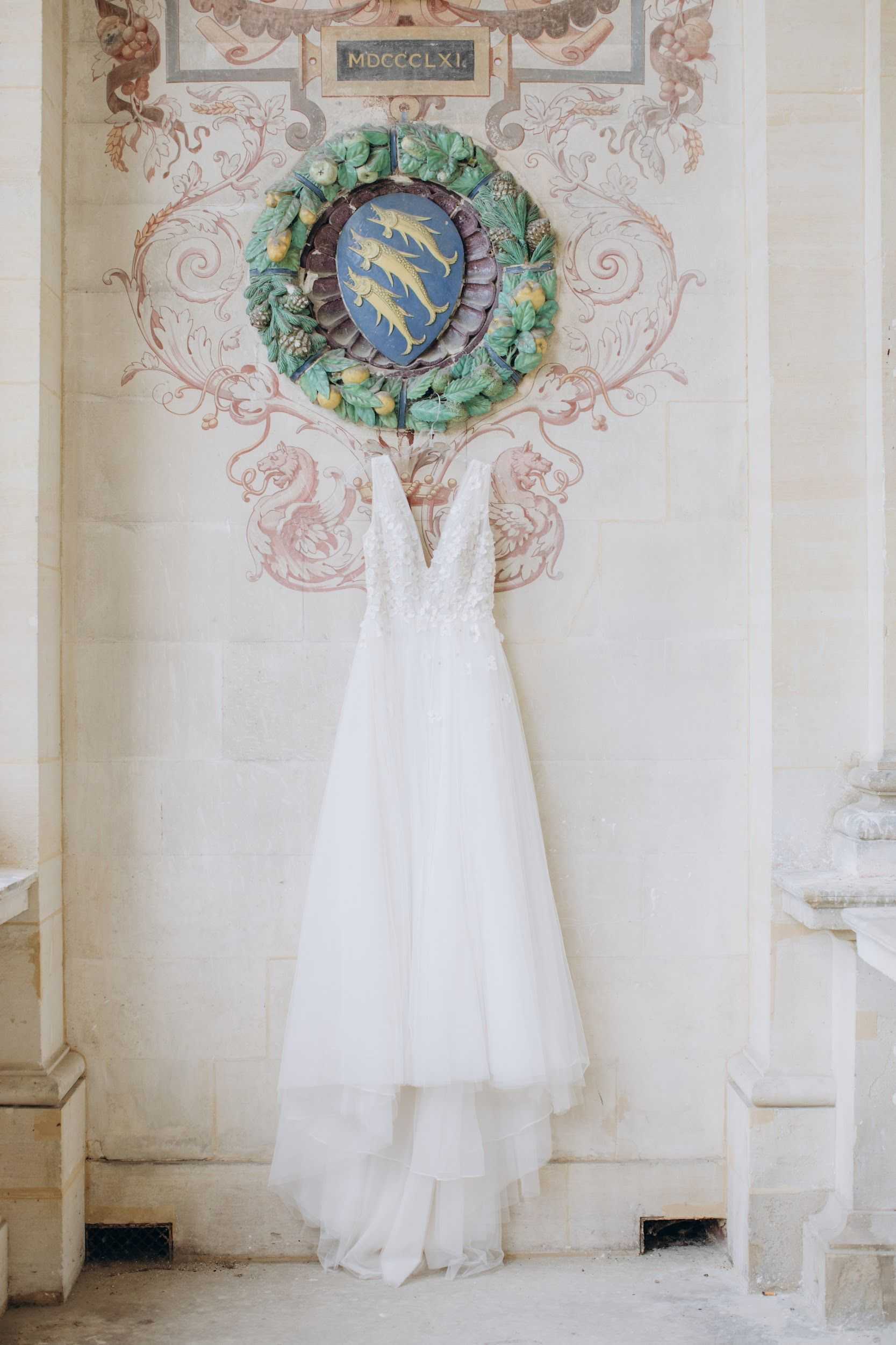 A detail shot of a white bridal gown hanging against an ornate stone wall of what appears to be a French chateau or historic venue. The dress features a lace-appliquéd V-neck bodice and a layered tulle skirt, and is suspended from a decorative ceramic wall medallion depicting three golden fish on a blue heraldic shield, surrounded by a wreath of sculpted green leaves and fruit, with the Roman numeral date MDCCCLXI inscribed above. The surrounding wall is pale limestone with painted pink and grey scrollwork and griffon motifs in a classic French architectural style. The composition is a vertical full-length detail shot centered on the dress against the architectural backdrop. Potential venue feature image.