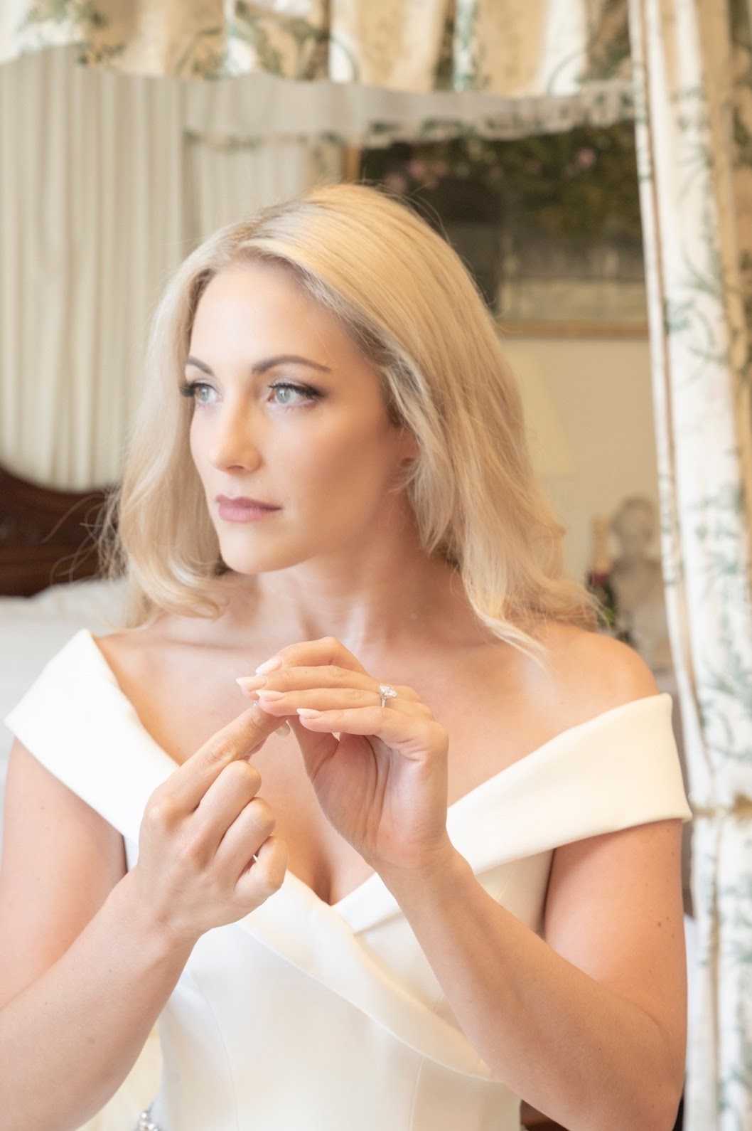 A bride is photographed indoors during a getting-ready moment, gazing off to one side with her hands clasped in front of her chest, drawing attention to her diamond engagement ring. She wears a structured off-the-shoulder ivory wedding gown with a clean, modern neckline and has her blonde hair styled in loose waves. The background shows a traditionally decorated bedroom with floral and striped curtains and a four-poster bed, suggesting a chateau or manor house setting. This is a close-up portrait shot with soft, warm lighting and a shallow depth of field that keeps the focus on the bride's face and hands.
