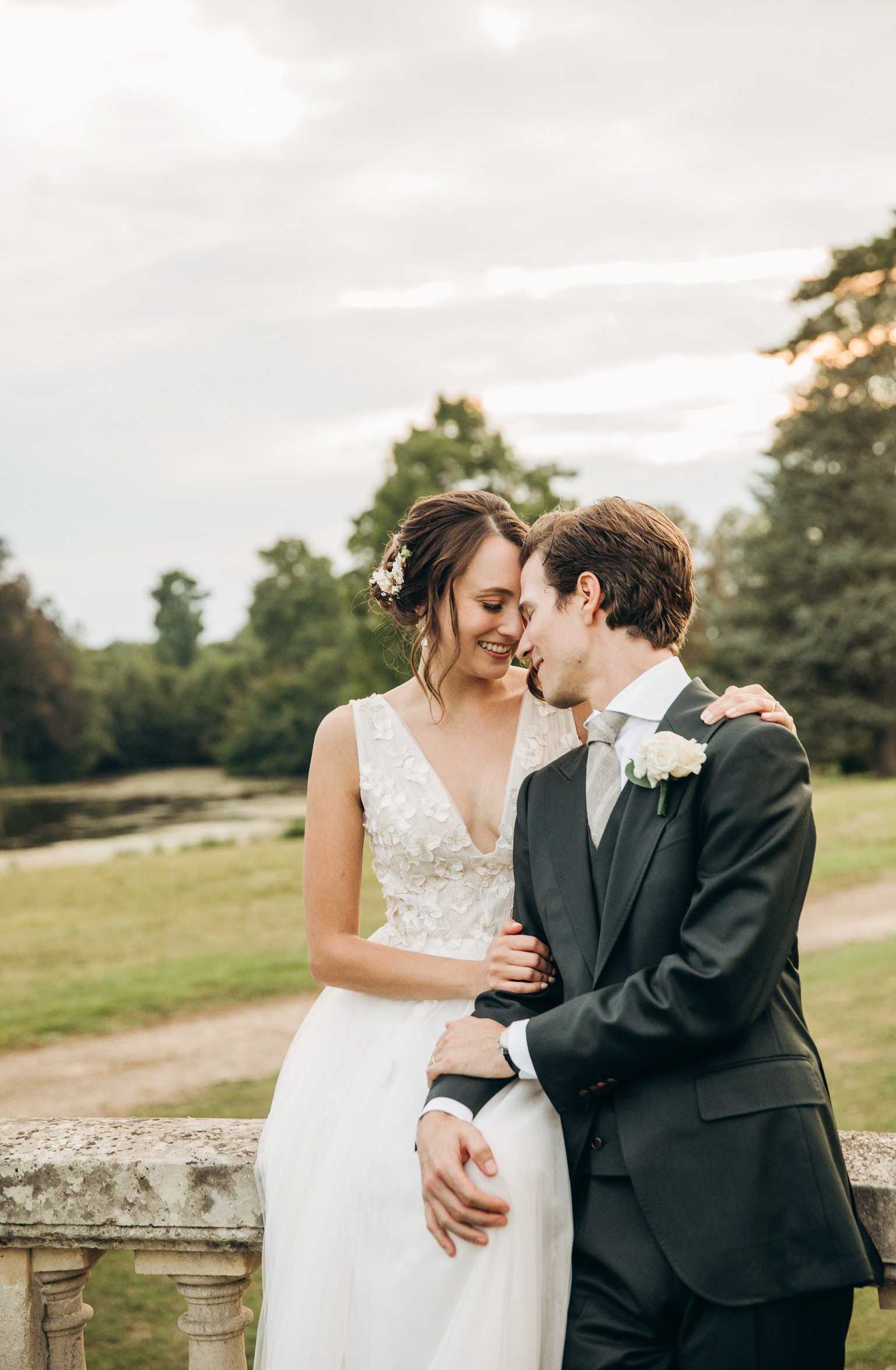 An outdoor couple portrait taken at golden hour, with the bride and groom standing close together against a stone balustrade, likely on the terrace of a chateau or manor estate. The bride wears an ivory sleeveless V-neck gown with three-dimensional floral appliqué detailing on the bodice, her hair styled in a loose updo with small white floral hair accessories. The groom wears a dark charcoal suit with a silver tie and a white rose boutonniere. The composition is a mid-length portrait shot with a softly blurred parkland background, giving the image a classic, romantic feel with warm evening light.