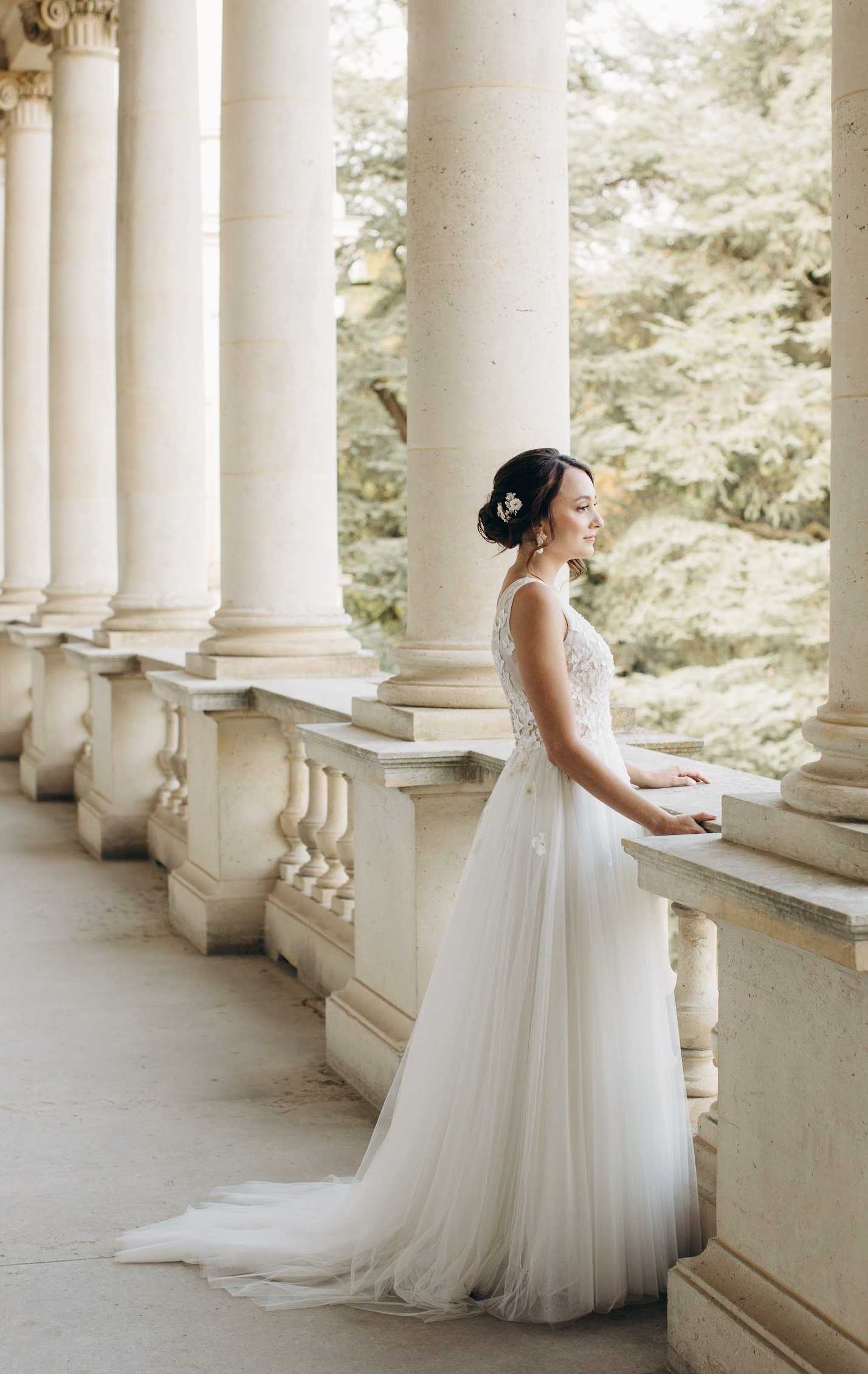 A bridal portrait taken on a grand stone colonnade, with the bride standing in profile and resting her hand on a carved balustrade. She wears a floor-length ivory gown with a lace-appliqué bodice and a soft tulle skirt with a flowing train, paired with a delicate floral hair comb and drop earrings, her dark hair styled in an updo. The setting features a row of tall classical columns with Corinthian capitals extending into the background, creating strong leading lines. The shot is a full-length portrait with a classic, formal styling aesthetic. Potential venue feature image.