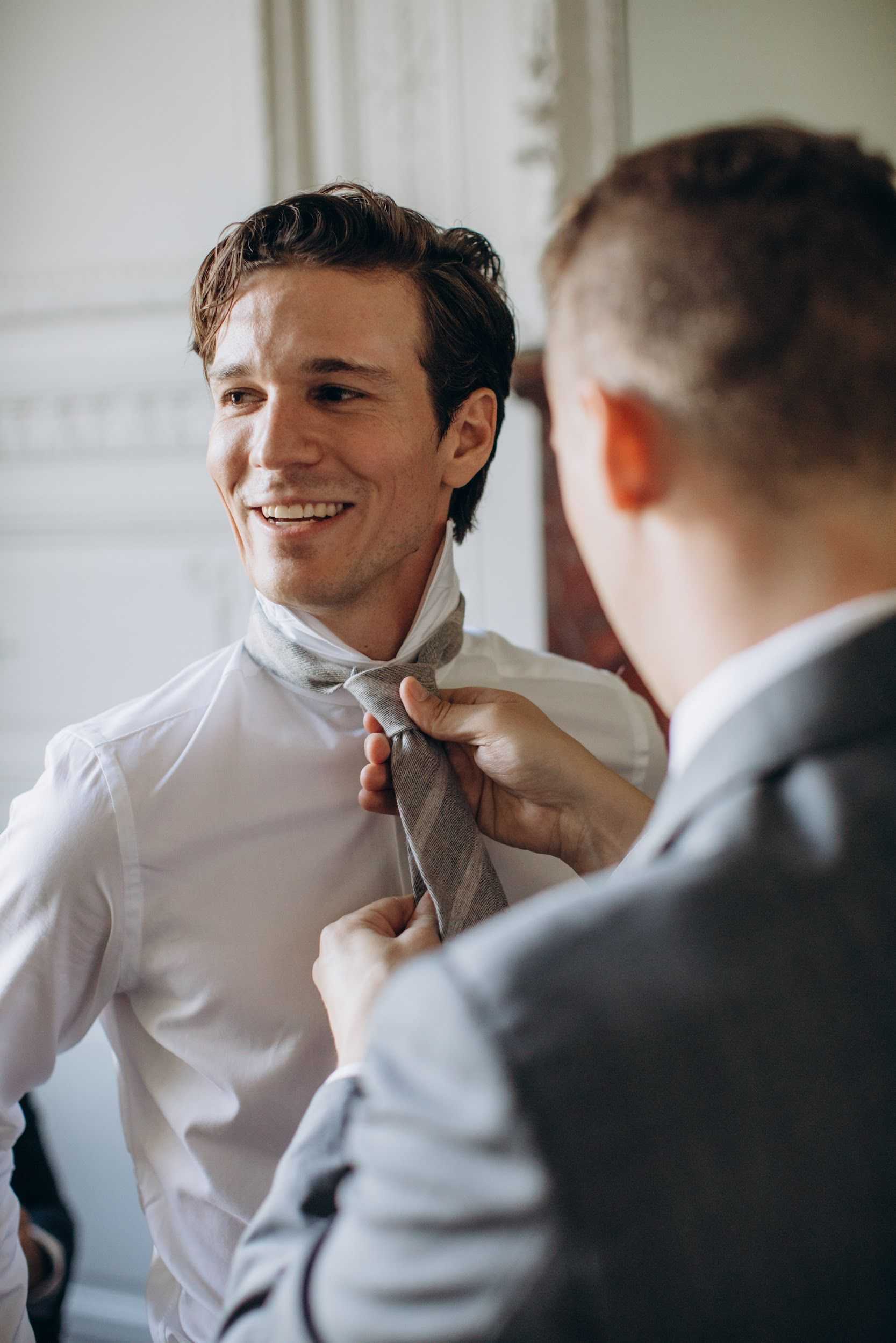 A getting-ready moment captured indoors, likely in a chateau or formal venue with white ornate molding visible in the background. A smiling groom in a white dress shirt is having a grey textured tie adjusted by a second man wearing a light grey suit jacket, shot from behind over his shoulder. The composition is a close-up portrait with soft natural window light illuminating the groom's face, creating a warm, candid feel with a shallow depth of field that blurs the foreground figure.