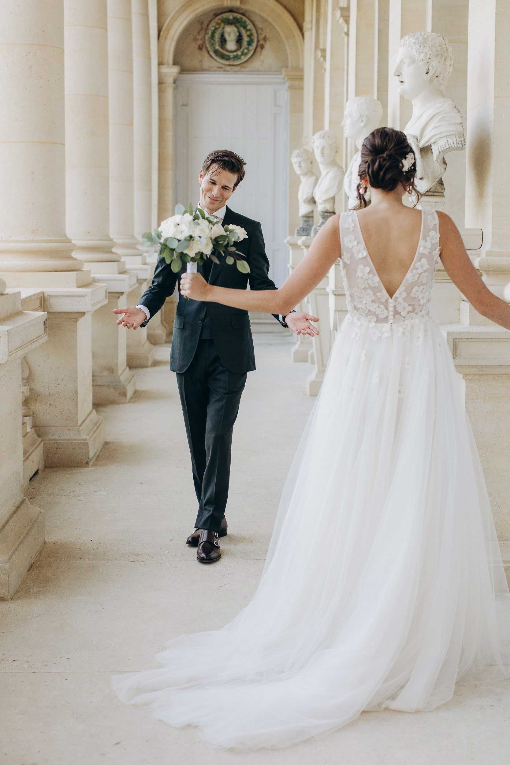 A couple's portrait capturing what appears to be a first look or playful reunion moment in a grand classical interior gallery lined with stone columns, arched ceilings, and a row of white marble busts on pedestals. The groom, wearing a dark navy suit with black dress shoes and a white boutonnière, walks toward the bride with arms open wide and a broad smile, holding the bridal bouquet of white blooms and eucalyptus greenery in one hand. The bride faces away from the camera, showing the back of her white ball gown featuring a deep V-back with floral appliqué detailing on the bodice and a long tulle train pooling on the floor; her dark hair is styled in an updo with a floral hair accessory. The composition is a medium full-length portrait shot from behind the bride, creating depth down the colonnaded corridor.