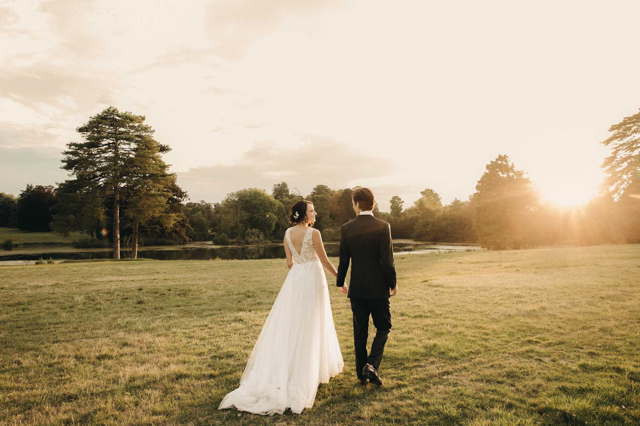 A couple portrait taken outdoors during golden hour, with the bride and groom walking hand-in-hand across an open lawn toward a small lake or pond in the background. The bride wears a white A-line gown with a lace appliqué bodice, a deep V-back, and a flowing tulle skirt with a short train; her dark hair is styled up with a small floral hair accessory. The groom wears a classic black suit with a white dress shirt. The bride turns her head back slightly with a smile while the groom faces forward. The shot is a wide, full-length portrait taken from behind, bathed in warm amber sunlight flaring in from the right side of the frame.
