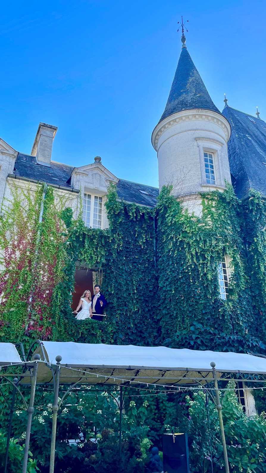 A bride and groom pose on a first-floor balcony doorway of a French chateau, shot from ground level looking up in a wide-angle perspective. The bride wears a white off-the-shoulder gown and the groom wears a navy blue suit with a boutonniere. The chateau facade is heavily covered in green and burgundy climbing vines, with a prominent round turret topped by a dark slate conical roof visible to the right. In the foreground, a white-canopied iron pergola structure with string lights is partially visible, along with flowering garden plants below. Potential venue feature image.