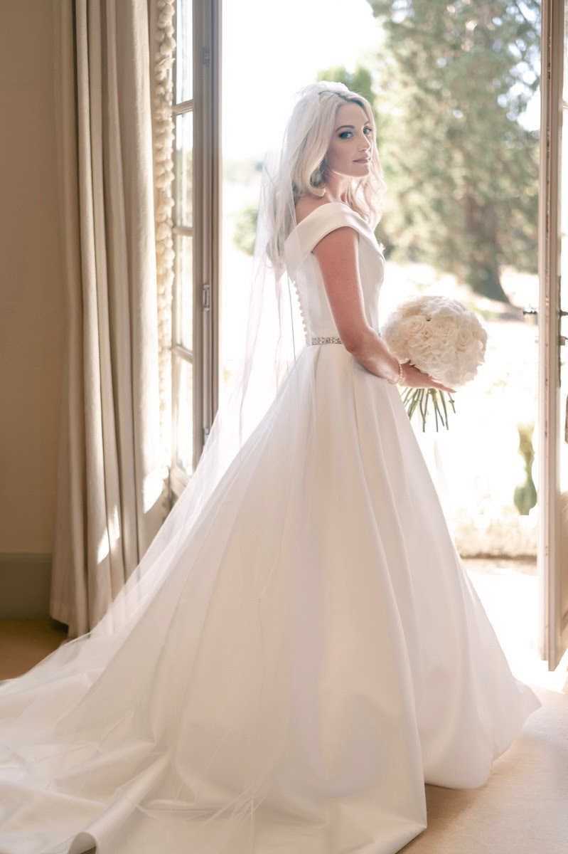 A bridal portrait of a single bride standing in an open set of French doors, looking back over her shoulder toward the camera. She wears a white off-the-shoulder ball gown with a fitted bodice featuring a crystal-embellished belt, a long cathedral train, and a shoulder-length veil. She holds a compact round bouquet of ivory peonies or garden roses. The interior setting shows warm beige floor-length curtains and warm-toned flooring, consistent with a chateau or manor house interior, while the open doors reveal a sunny outdoor landscape. The shot is a full-length portrait with natural backlight creating a soft, bright contrast between the interior and exterior.