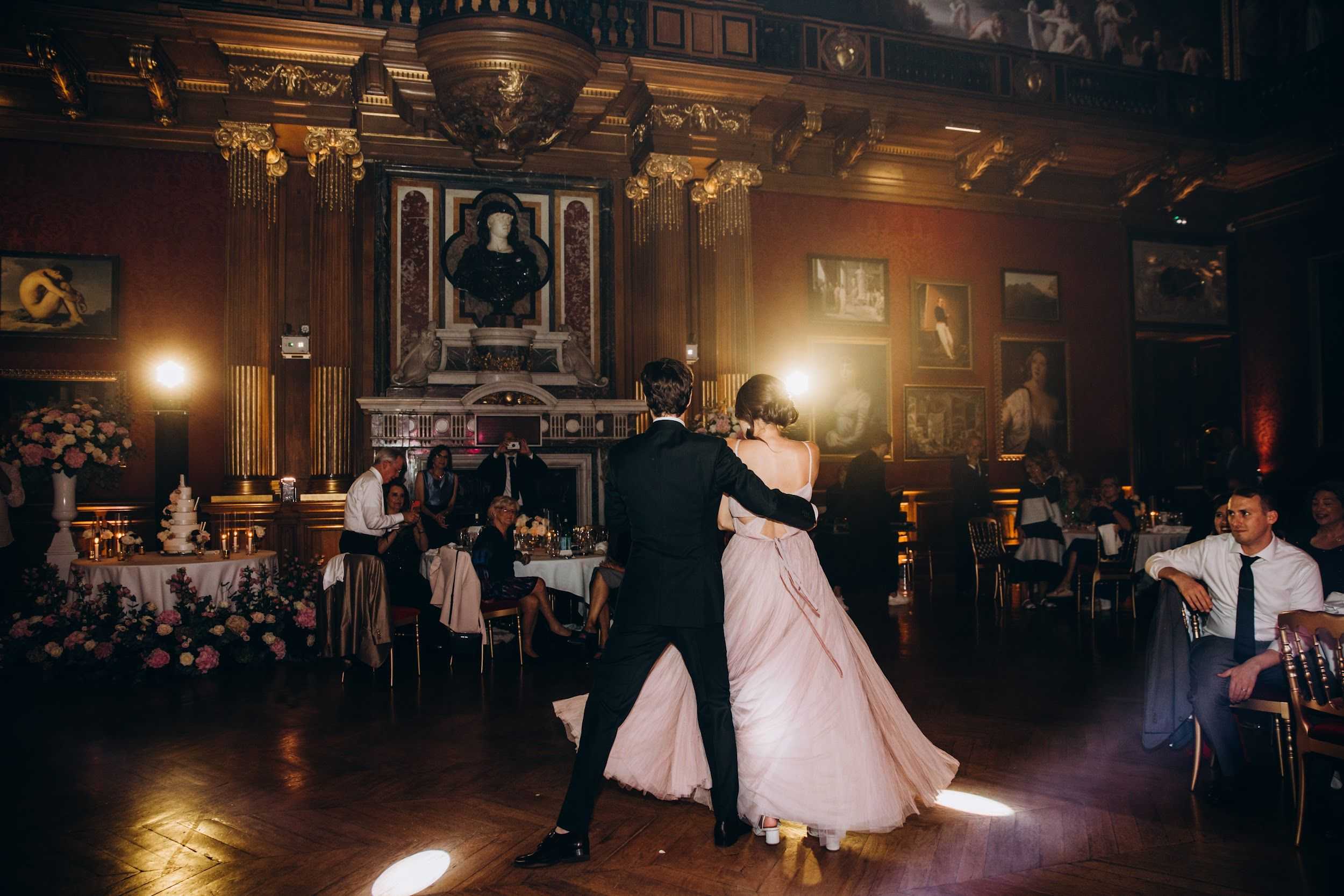 A couple shares their first dance on a parquet wood floor in an ornate ballroom with dark wood paneling, gilded Corinthian columns, large oil paintings on the walls, and an elaborate marble fireplace topped with a classical bust as the central backdrop. The bride wears a flowing blush pink tulle gown with a low open back and spaghetti straps, while the groom is in a dark navy or black suit; the two are photographed from behind in a wide shot with warm spotlight illumination. Seated guests line the perimeter of the dance floor watching the dance, with round tables dressed in white linens, gold candleholders, and floral centerpieces visible in the background, along with a multi-tiered white wedding cake on a separate table adorned with pink and blush floral arrangements at its base. The overall decor palette is blush, pink, and gold against the rich dark tones of the room, creating a classic formal reception atmosphere.