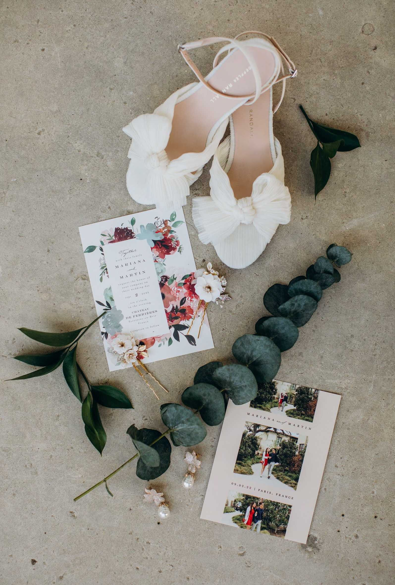 A flat lay detail shot of bridal accessories and stationery arranged on a concrete surface. The composition includes a pair of white strappy heeled sandals with ruffled bow detail (Loeffler Randall style), a wedding invitation suite featuring watercolor burgundy and blush florals with greenery for Mariana and Martin at Chateau de Fereires, France, a save-the-date card with couple photos dated 09.02.22 in Paris, France, gold hair pins with small ivory floral accents, pearl and crystal drop earrings, and stems of eucalyptus and dark green foliage used as styling props. The stationery palette is white and cream with deep red, blush, and sage green watercolor florals, consistent with a classic romantic styling theme.