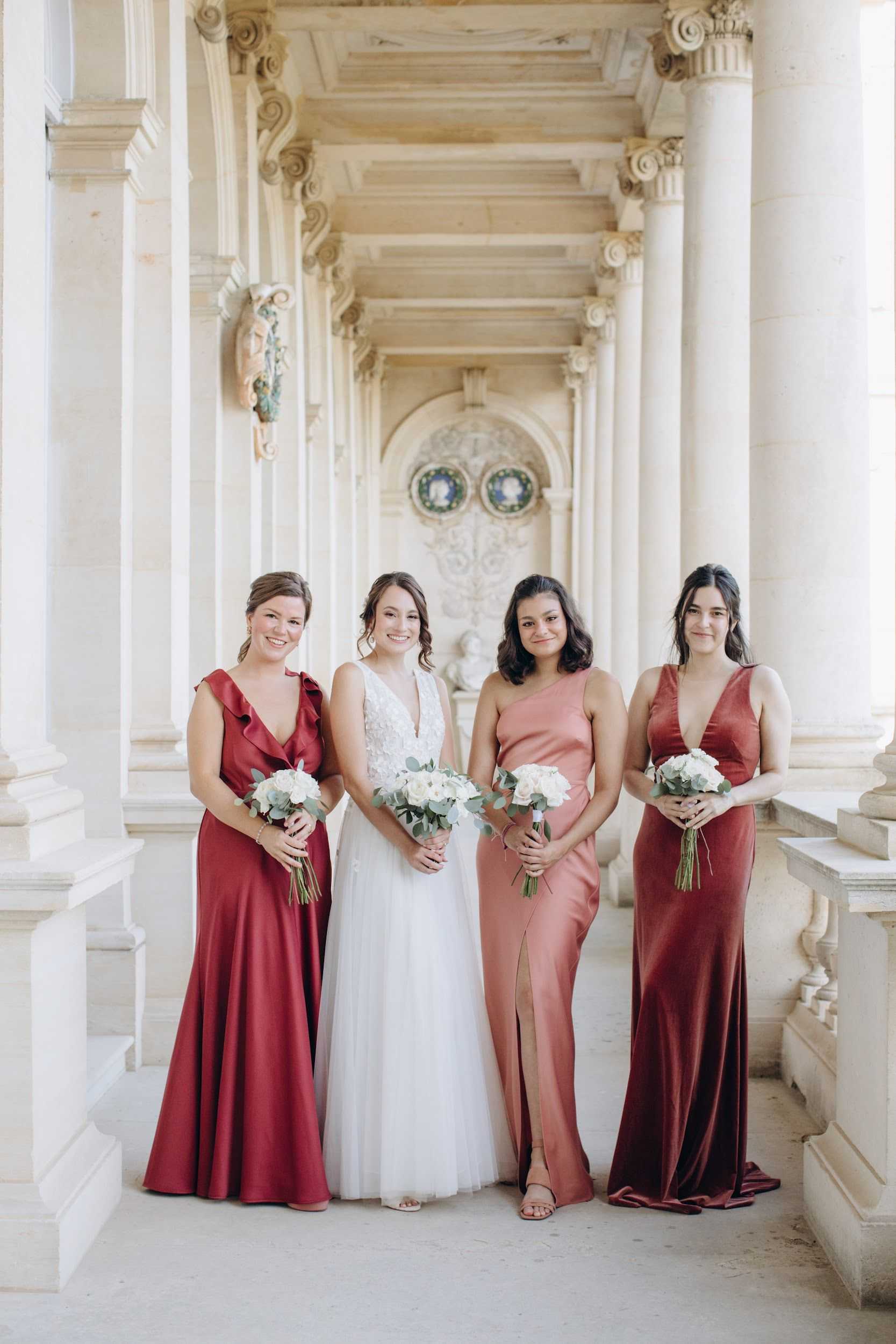 A bridal party portrait featuring the bride and three bridesmaids posing in a grand colonnaded gallery with ornate Corinthian columns, carved stone details, and decorative arched niches in the background, consistent with a classical French palace or museum setting. The bride wears a white deep V-neck lace and tulle gown, while two bridesmaids wear deep crimson satin floor-length dresses in slightly different styles, and one bridesmaid wears a dusty rose satin floor-length dress with a front slit. All four women hold small round bouquets of white roses and white blooms with eucalyptus greenery. The shot is a full-length group portrait taken in natural light with a bright, airy tone. Potential venue feature image.