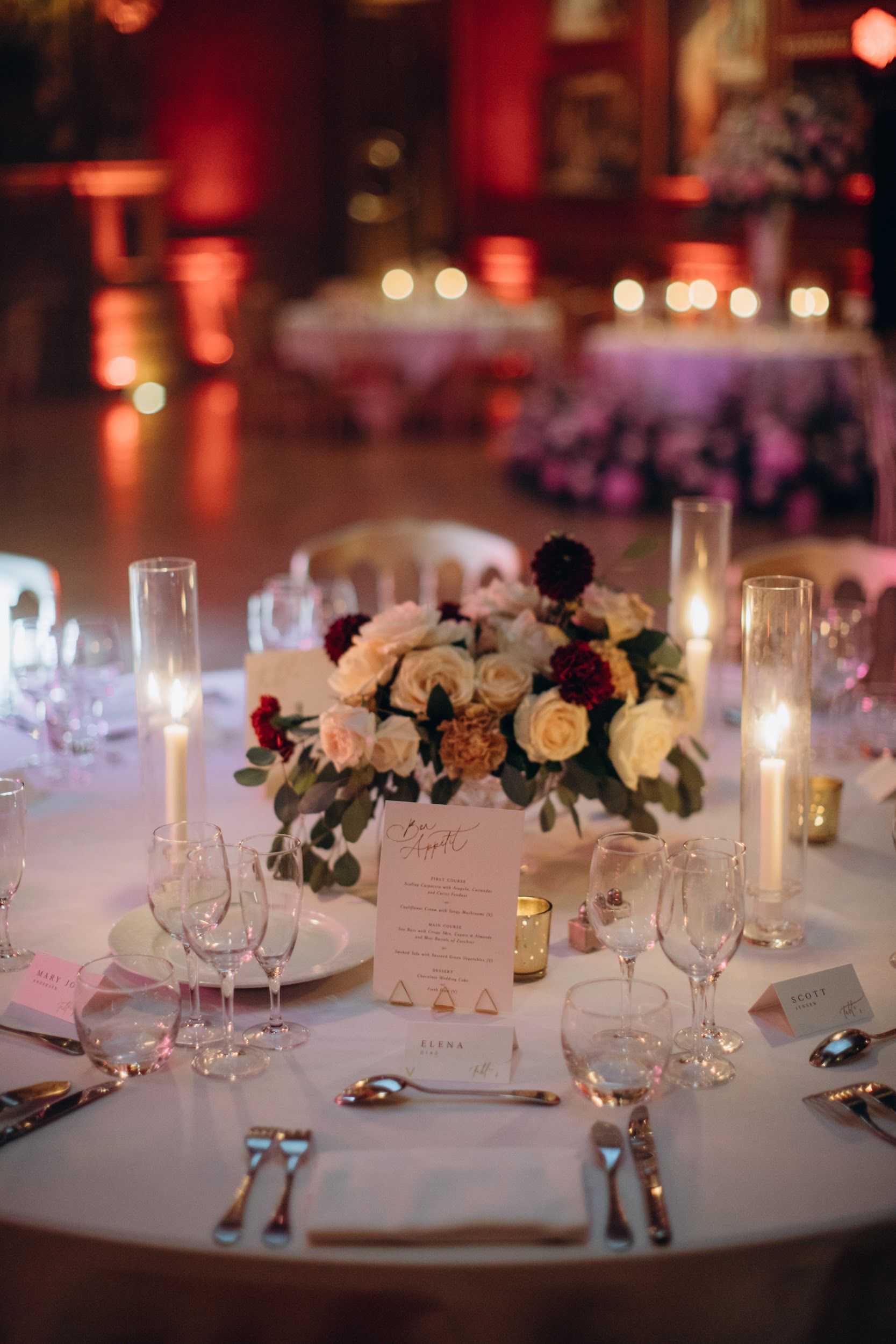Close-up detail shot of a wedding reception table setting in an indoor ballroom, lit with warm red and amber uplighting in the background. The round table is covered with a white linen and set with silver cutlery, clear wine glasses, white charger plates, and individual place cards with guest names including 'Elena' and 'Scott' visible. The low centerpiece features cream and blush roses, deep burgundy dahlias, mustard carnations, and eucalyptus foliage, surrounded by tall glass hurricane candle holders with white pillar candles and small gold votive candles. A blush-toned menu card with calligraphy script reading 'Bon Appétit' is propped against the centerpiece. In the soft-focus background, additional candlelit tables and warm red uplighting create a rich, moody atmosphere consistent with a classic or romantic reception style.