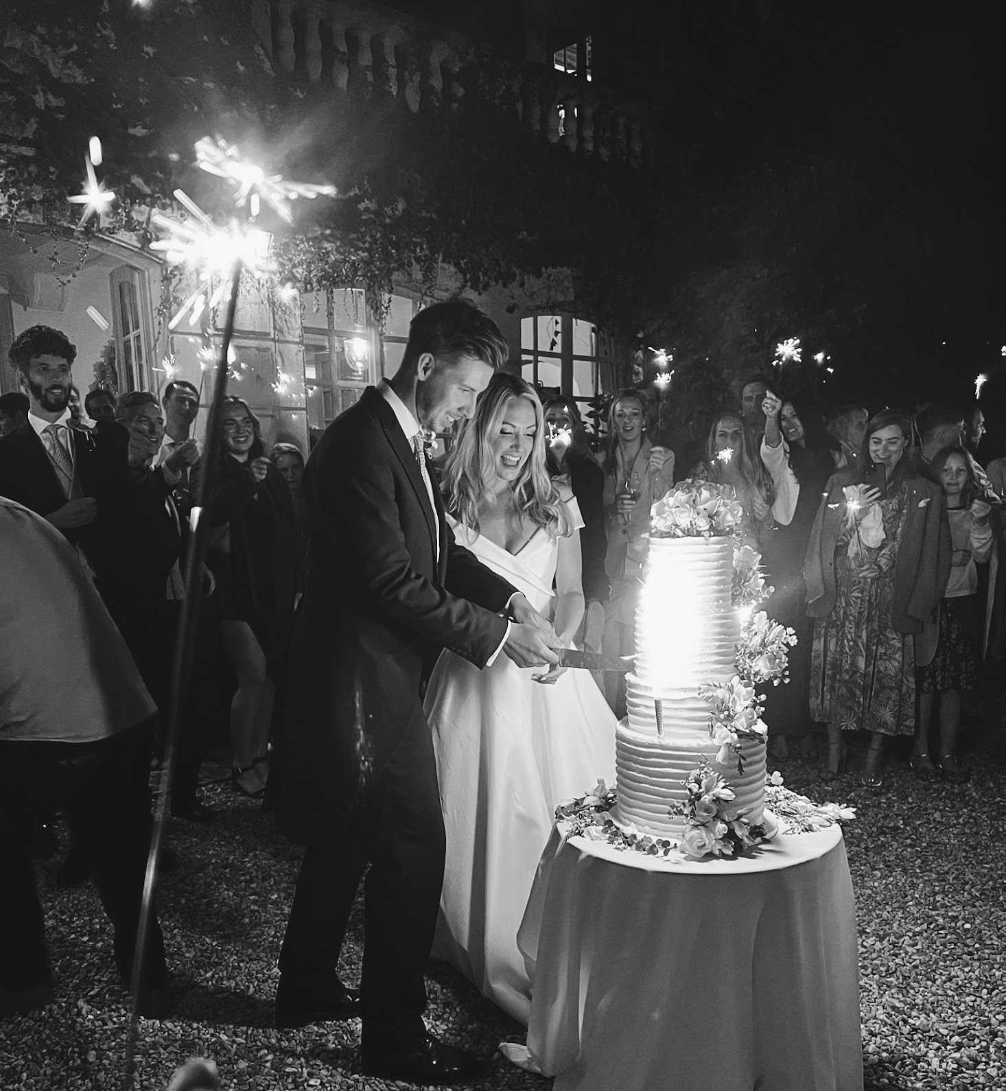 A black-and-white nighttime outdoor cake-cutting moment in front of what appears to be a chateau or manor house facade with lit windows. The bride, in a V-neck sleeveless gown, and the groom, in a dark suit, cut together into a three-tier horizontally ribbed buttercream cake decorated with florals at the base and top, placed on a satin-draped round table. Approximately 30 guests surround the couple in a semicircle, many holding large lit sparklers that create bright streaks and strong contrast in the black-and-white tones. The high-contrast image captures the sparkler light illuminating the couple's laughing expressions, with the gravel courtyard ground visible in the foreground. Wide shot with the venue building prominently visible in the background.
