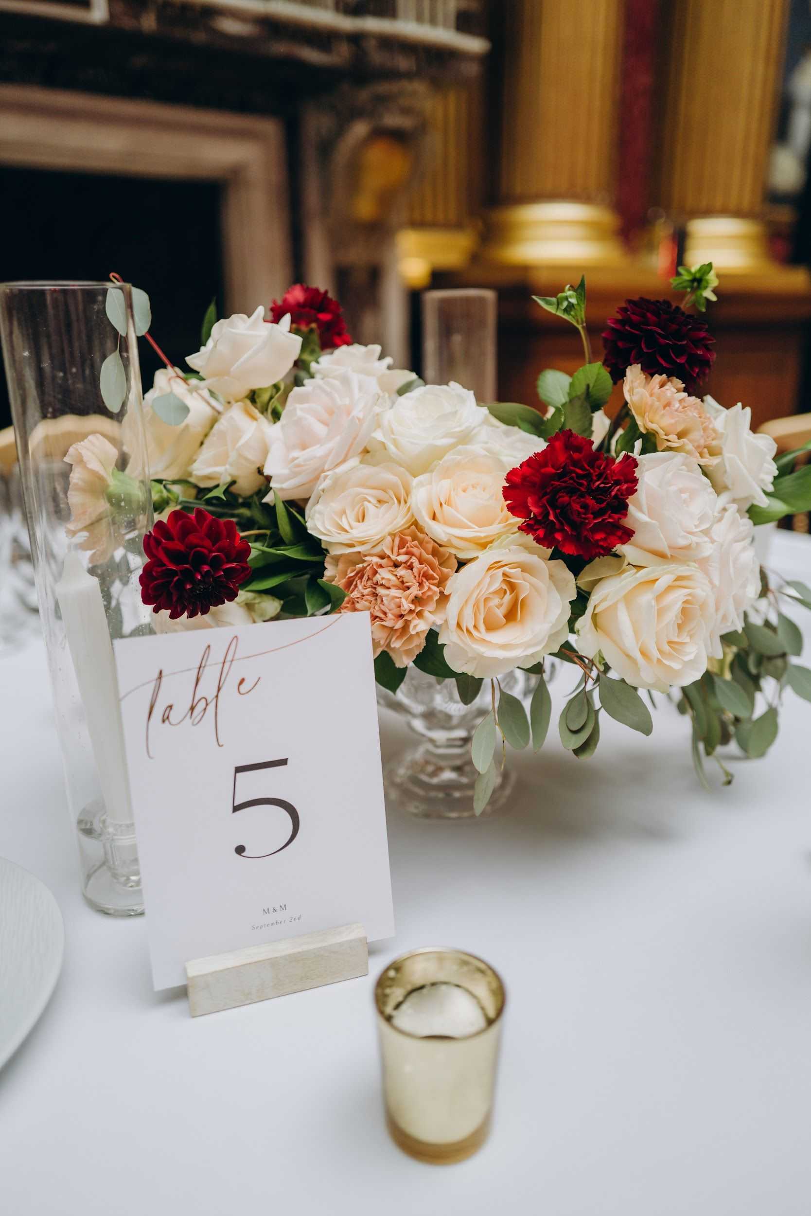 Close-up detail shot of a wedding reception table centerpiece in an indoor ballroom or formal hall setting, with ornate gold architectural details and a fireplace mantel visible in the background. The low floral arrangement sits in a clear glass compote vase and features cream and blush garden roses, deep crimson dahlias, red carnations, peach carnations, and eucalyptus greenery. A white table number card reading 'Table 5' with burgundy script lettering is displayed in a gold stand alongside a small gold votive candle holder and a tall glass cylinder vase, all on a white linen tablecloth. The overall decor palette is cream, blush, and deep red with gold accents, reflecting a classic, formal styling.