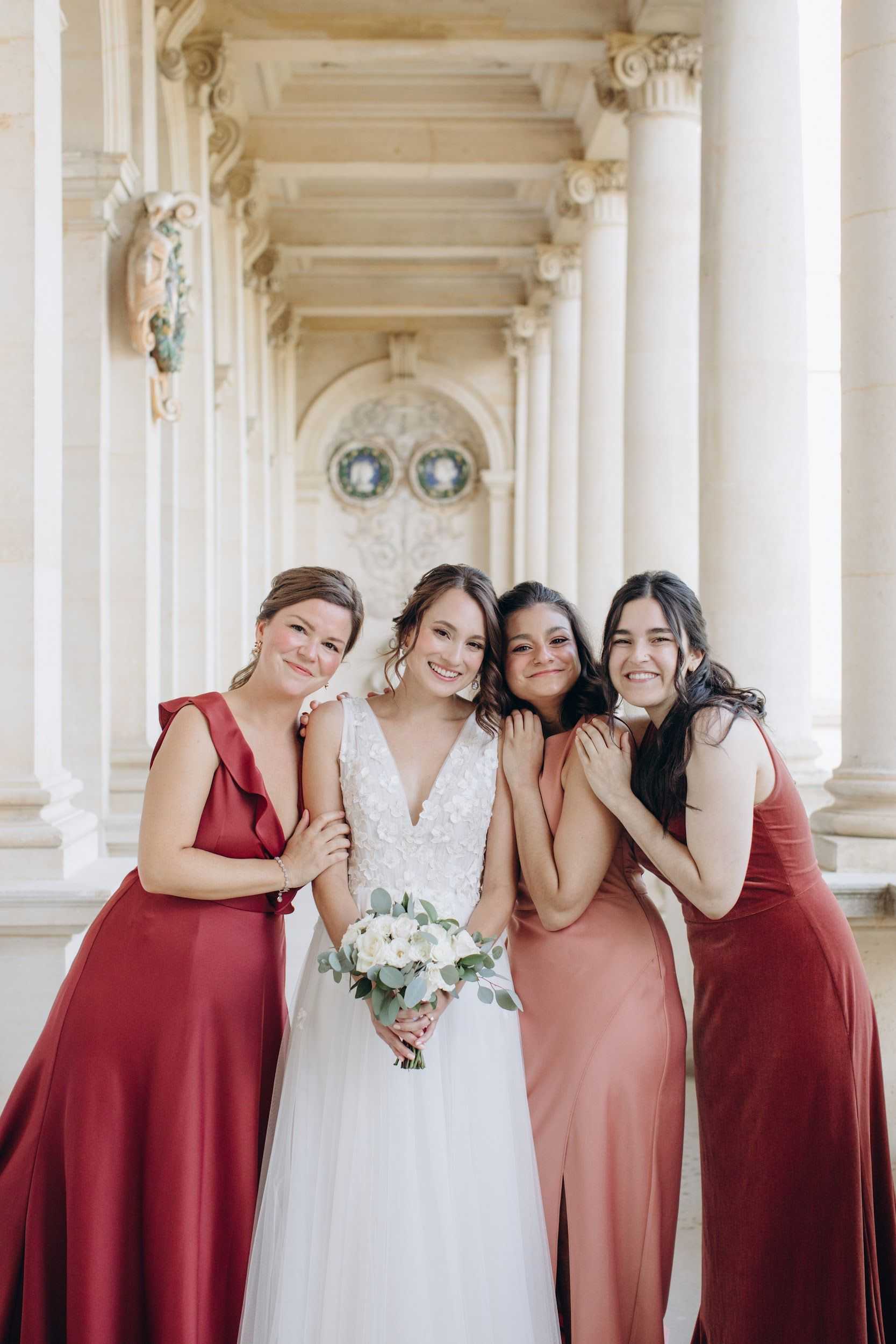 A bridal party portrait featuring the bride and three bridesmaids posed together in a grand colonnaded hallway with tall classical columns, ornate Corinthian capitals, and decorative carved stone details in the background. The bride wears a white V-neck gown with a floral appliqué bodice and holds a compact bouquet of white ranunculus and eucalyptus; the bridesmaids wear floor-length gowns in mixed shades of deep crimson and dusty rose/terracotta, with varying necklines including a ruffled V-neck and a simple halter style. The group is relaxed and candid, with the bridesmaids leaning in toward the bride and smiling warmly. The shot is a mid-length portrait taken in natural light within what appears to be a formal French palace or château arcade.