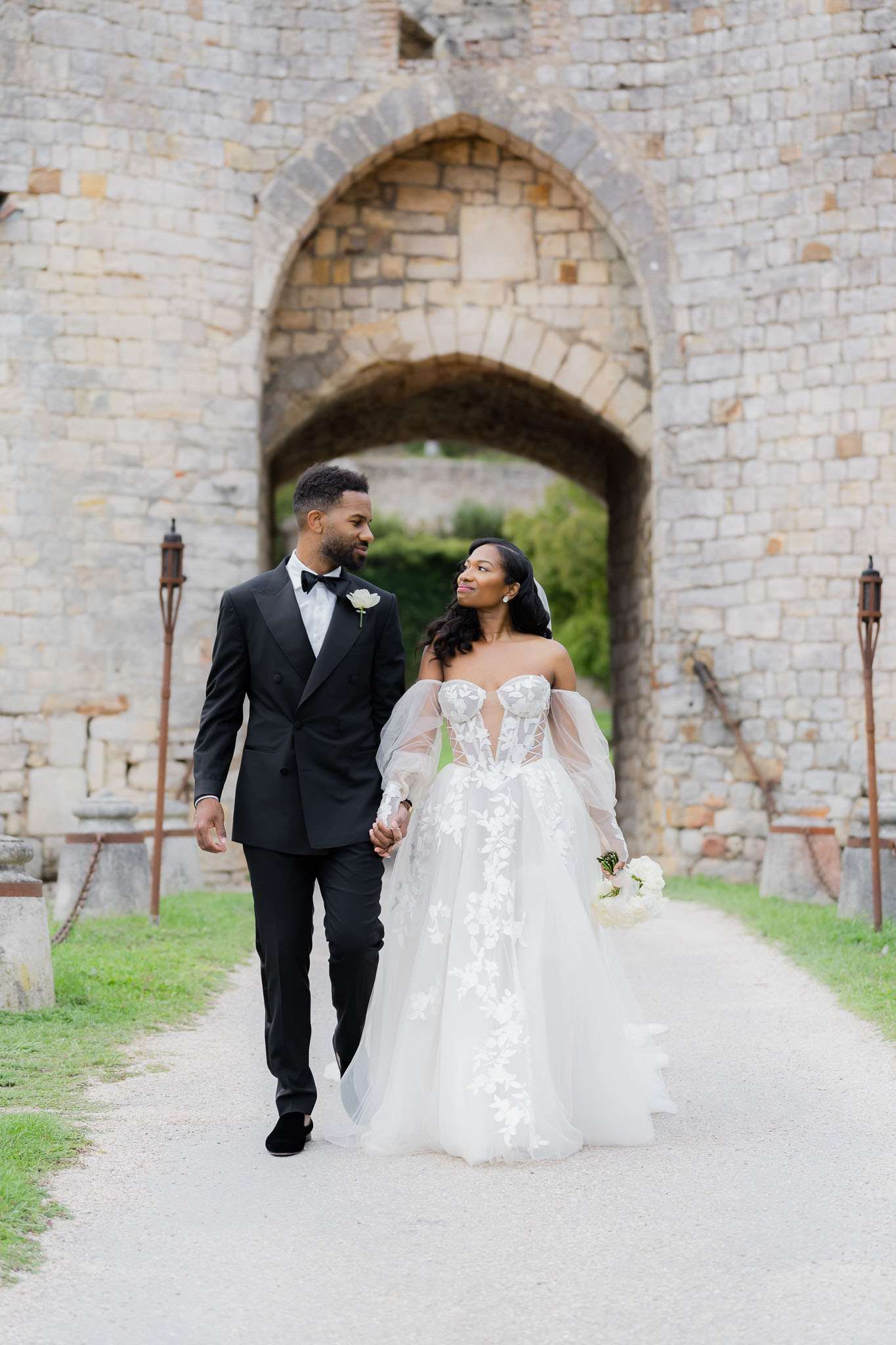 A couple portrait taken outdoors at a historic château or castle, with the bride and groom walking hand-in-hand along a gravel path toward the camera. A large Gothic-arched stone gateway serves as the backdrop. The groom wears a black double-breasted tuxedo with a bow tie and a white floral boutonniere. The bride wears an off-the-shoulder white ballgown with a sheer lace-appliquéd bodice featuring floral detailing, voluminous tulle skirt, and sheer puff sleeves; she carries a bouquet of white peonies or ranunculus. The styling is classic and formal. The shot is a full-length portrait with the archway centered in soft focus behind the couple.