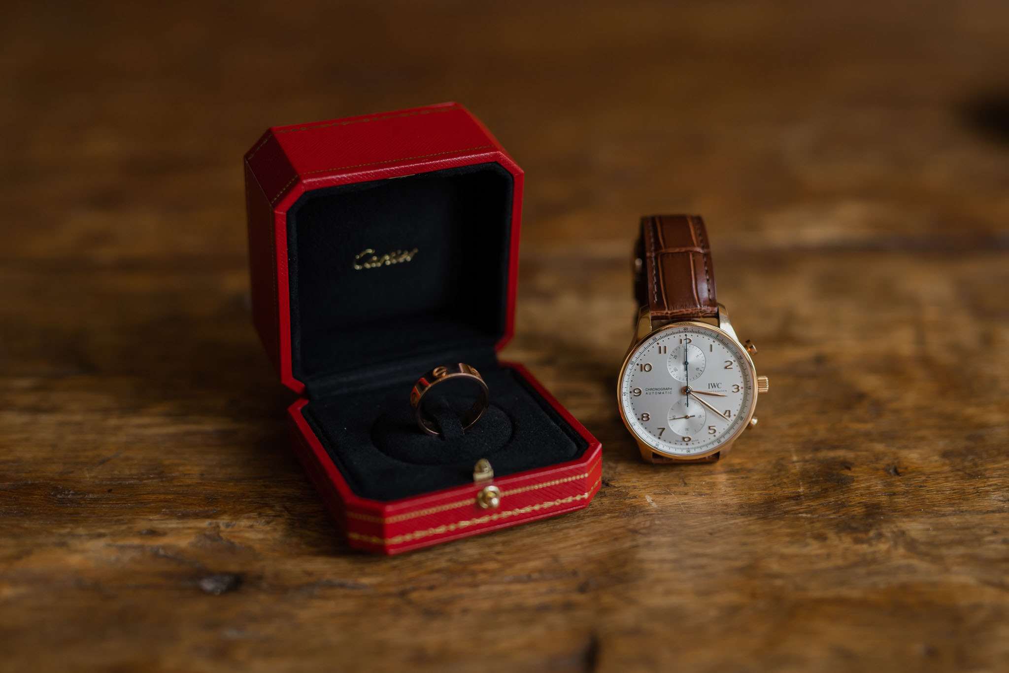 A close-up detail shot of groom's accessories arranged on a worn wooden surface. A rose gold Cartier wedding band sits inside its open red Cartier box with black velvet interior, positioned next to an IWC Portugieser Chronograph automatic watch featuring a white dial, gold-tone case, and brown crocodile-embossed leather strap. The warm, dark wooden background creates a rich contrast against the red box and metallic accessories.