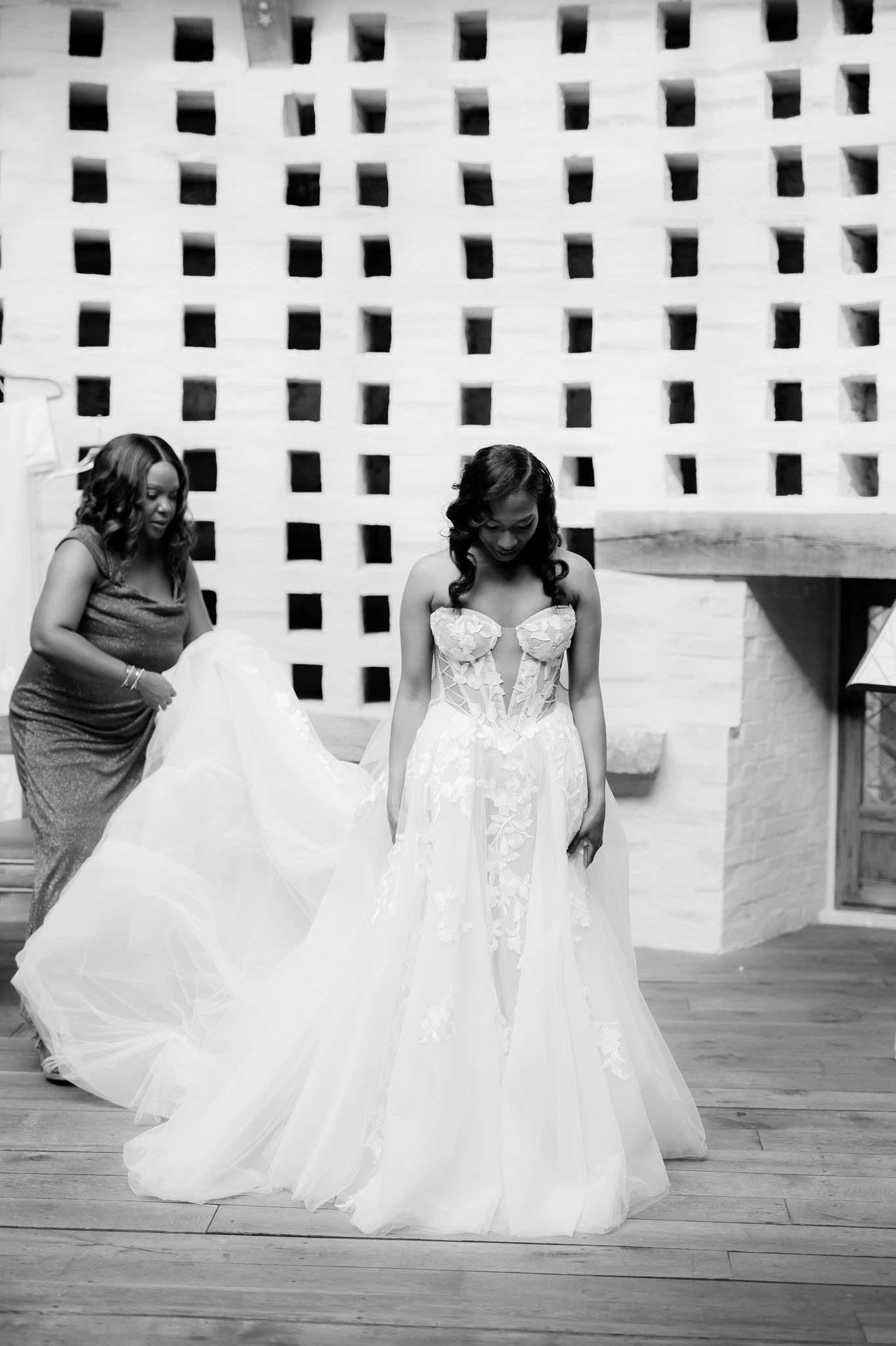 This black-and-white portrait captures a getting-ready moment on a wooden deck, where a bride stands looking downward while a second woman in a floor-length fitted dress arranges and lifts the train of the bridal gown. The bride wears a strapless ballgown with a structured floral appliqué bodice and a full layered tulle skirt, her hair styled in loose curls. The background features a geometric lattice or breeze-block wall with a light, bright tone that contrasts against the darker mid-tones of the figures. The composition is a medium full-length shot with moderate depth of field, keeping both women in focus.