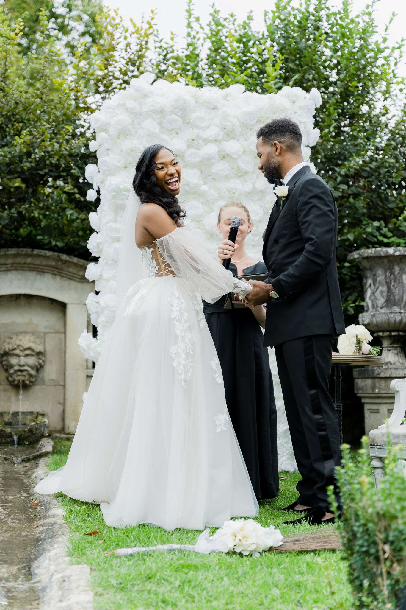 An outdoor wedding ceremony captured in a medium portrait shot, showing a bride and groom exchanging vows while laughing and holding hands, with an officiant standing between them holding a microphone. The ceremony takes place in a formal garden setting with a stone fountain featuring a carved face mask visible to the left and ornamental stone urns to the right. The backdrop is a large freestanding floral wall panel densely covered in white roses. The bride wears a white off-the-shoulder ballgown with a lace-appliquéd corset bodice, sheer long sleeves, and a full tulle skirt, with a cathedral-length veil. The groom wears a black tuxedo with a white boutonniere and black bow tie. A small cluster of white and cream flowers rests on the ground near the couple's feet, and a side table holds additional white and cream floral arrangements. The overall decor palette is white and black with a classic, formal aesthetic.