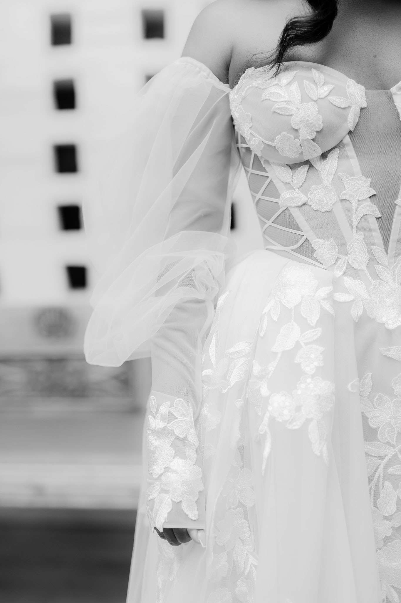 This is a black-and-white close-up portrait of a bride from the neck down, focusing on the bodice and sleeves of her wedding gown. The dress features a corset-style lace-up back with sheer tulle panels and intricate floral and leaf appliqué embroidery across the bodice, sleeves, and skirt. The sleeves are detachable off-the-shoulder puff sleeves in sheer tulle, adding a romantic, fashion-forward design element. The background is softly blurred, revealing what appears to be a light-colored architectural facade with evenly spaced rectangular openings.