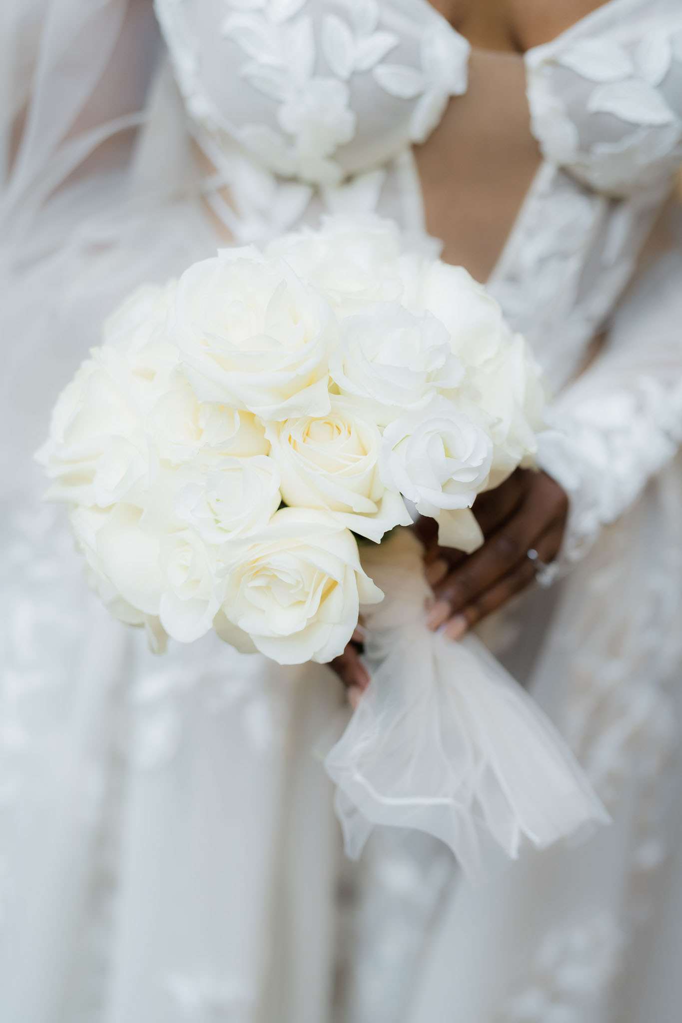 Close-up detail shot of a bride holding a tightly gathered round bouquet composed entirely of cream-white roses, wrapped with a sheer white organza ribbon at the stems. The bride is wearing a white long-sleeved gown with three-dimensional floral appliqué detailing on the bodice and sleeves, visible in the upper portion of the frame. The overall color palette is an all-white monochromatic scheme, and the styling is classic and minimal with no greenery or filler flowers in the bouquet.
