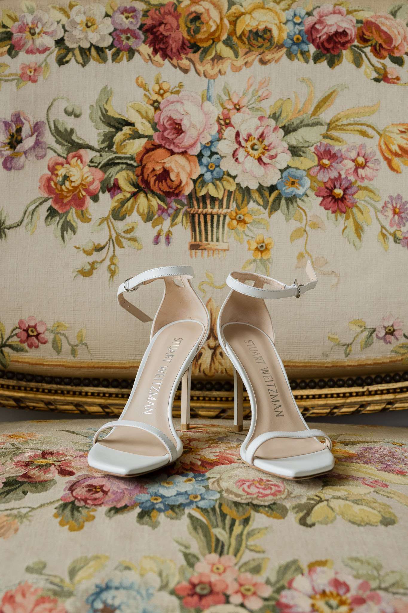 A close-up detail shot of a pair of white Stuart Weitzman strappy stiletto heels with ankle buckle closures and nude leather insoles, placed on an antique tapestry-upholstered chair or ottoman featuring a multicolored floral needlepoint pattern in pink, yellow, coral, blue, and ivory tones on a cream background with a gilt border. The shoes are positioned facing the camera, showing the brand name embossed on the insoles. The styling is minimal and classic, with the ornate vintage textile providing a decorative backdrop typical of a French chateau getting-ready setting.