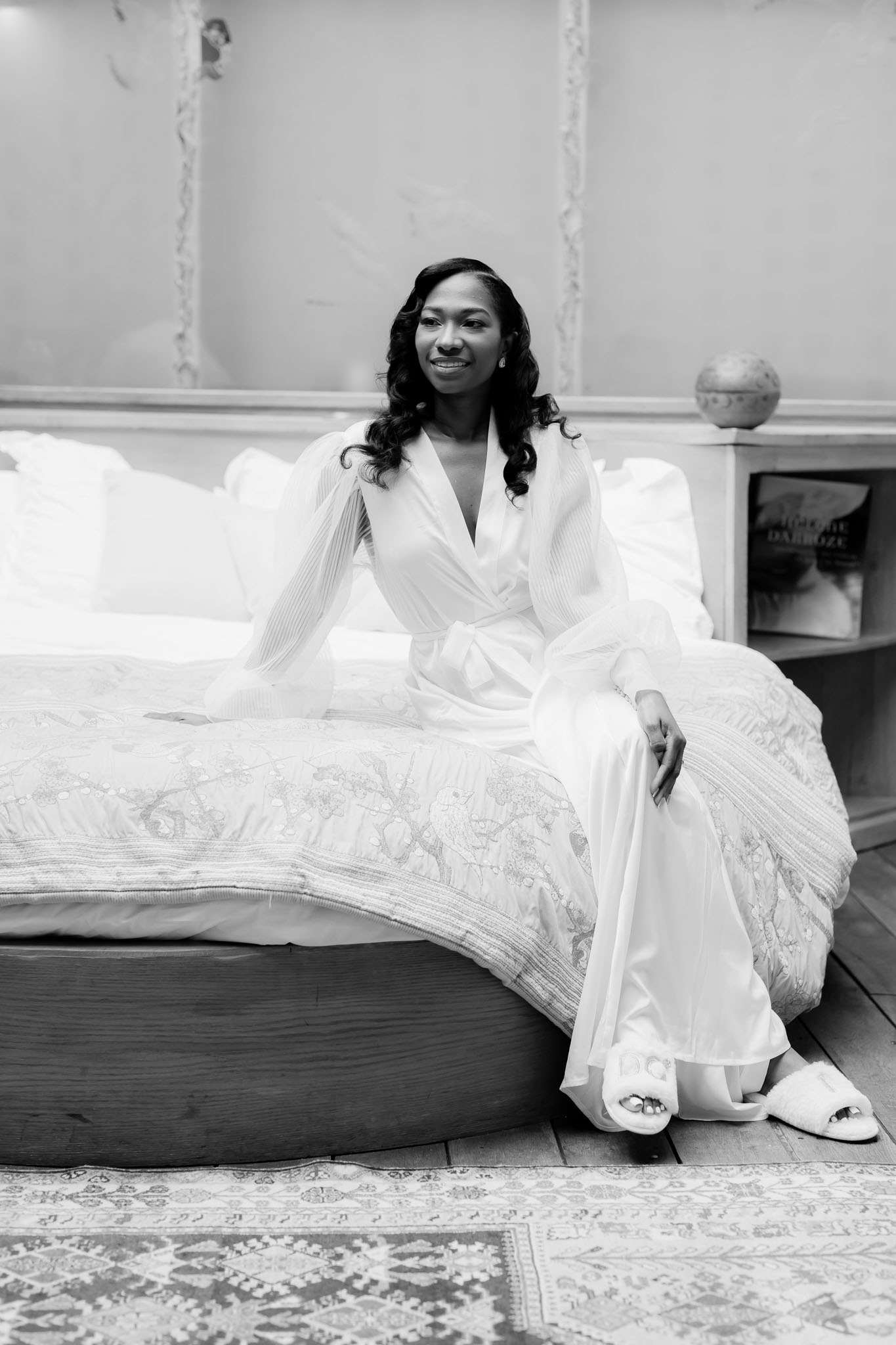 Black-and-white portrait of a bride during the getting-ready phase, seated on the edge of a large bed in an indoor bedroom setting. She wears a long white bridal robe with wide pleated sleeves and a wrap-tie waist, paired with fluffy white open-toe slippers, and her hair is styled in loose curls. The room features an ornate patterned bedspread, a patterned area rug, paneled walls with decorative molding, and a bedside table with a small decorative object and a book visible. The image is a full-length portrait shot with soft, even tones and moderate contrast, capturing a calm, composed moment before the ceremony.