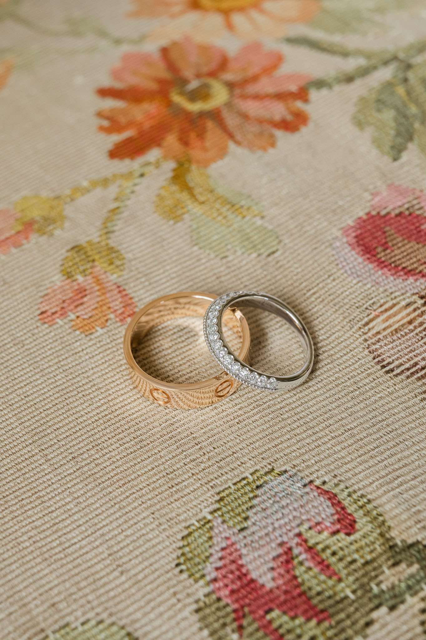 Close-up detail shot of two wedding bands placed side by side on a floral tapestry or embroidered fabric featuring coral, peach, and sage green botanical motifs on a cream background. The wider band is a rose gold Cartier Love ring with the signature screw detailing along its edge, while the second is a narrower white gold or platinum band fully set with pavé diamonds. The rings are leaning slightly against each other, styled without a ring box or pillow.