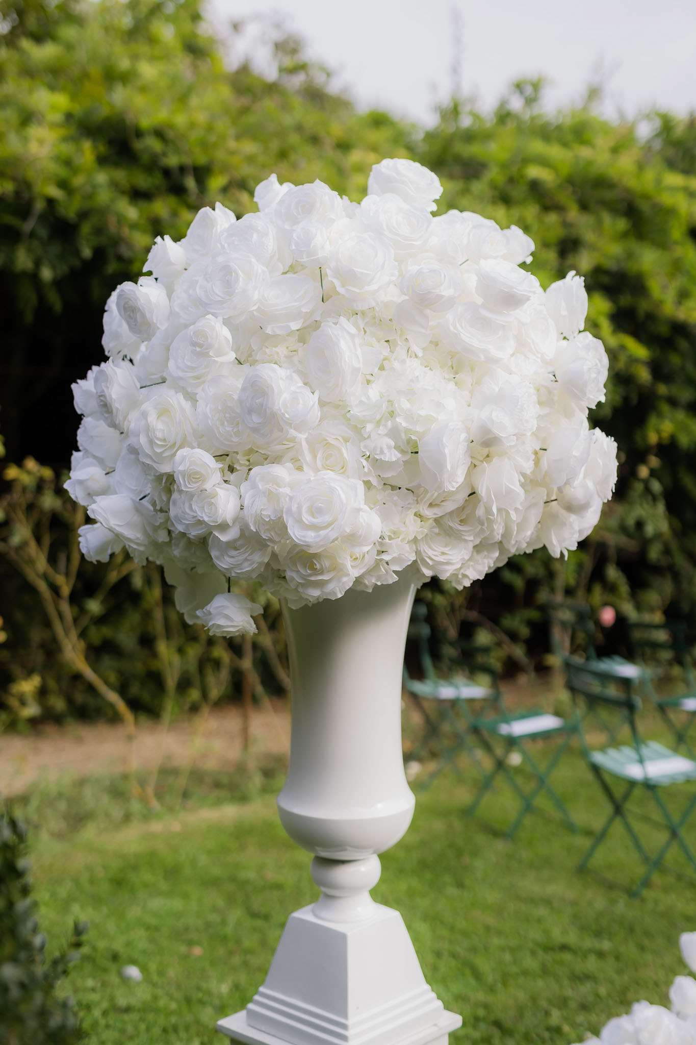 Close-up detail shot of a tall white pedestal urn holding a densely packed dome-shaped floral arrangement composed entirely of white roses, white ranunculus, and white hydrangeas. The arrangement is positioned outdoors in a garden setting, with green folding bistro chairs visible in the background suggesting a ceremony seating area. The all-white floral palette and classical urn-style pedestal reflect a formal, classic decor theme.
