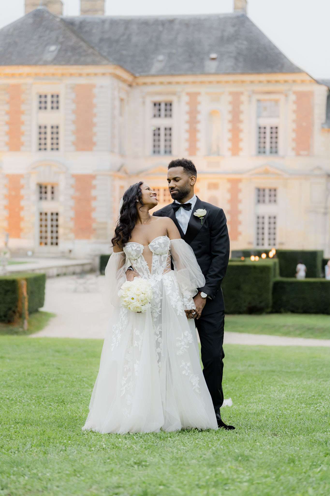 A couple portrait taken outdoors on the grounds of a French chateau, with the warm-toned pink and cream stone facade of a classic French manor house visible and softly out of focus in the background. The bride wears an off-shoulder white tulle ball gown with 3D floral appliqué detailing, a corset bodice, and detachable sheer puff sleeves, and carries a round bouquet of white peonies or ranunculus; the groom wears a black tuxedo with a black bow tie and a white floral boutonniere. The couple stands hand-in-hand on a lawn, the bride looking up at the groom with a smile while he looks down at her. The composition is a medium full-length portrait shot with trimmed hedgerows and warm string lights faintly visible in the background, suggesting an evening reception setting. Potential venue feature image.