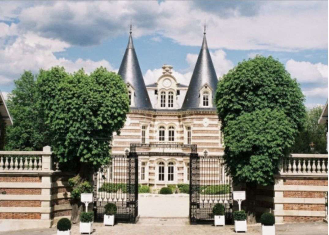 A wide exterior shot of a French château featuring two pointed slate-grey turrets, ornate red brick and cream stone striped façade, and arched windows with a central clock detail. The entrance is framed by wrought iron gates flanked by white planters with clipped boxwood topiaries, and a low brick balustrade runs along the front perimeter. No people are visible; the image focuses entirely on the architecture and grounds approach. Potential venue feature image.