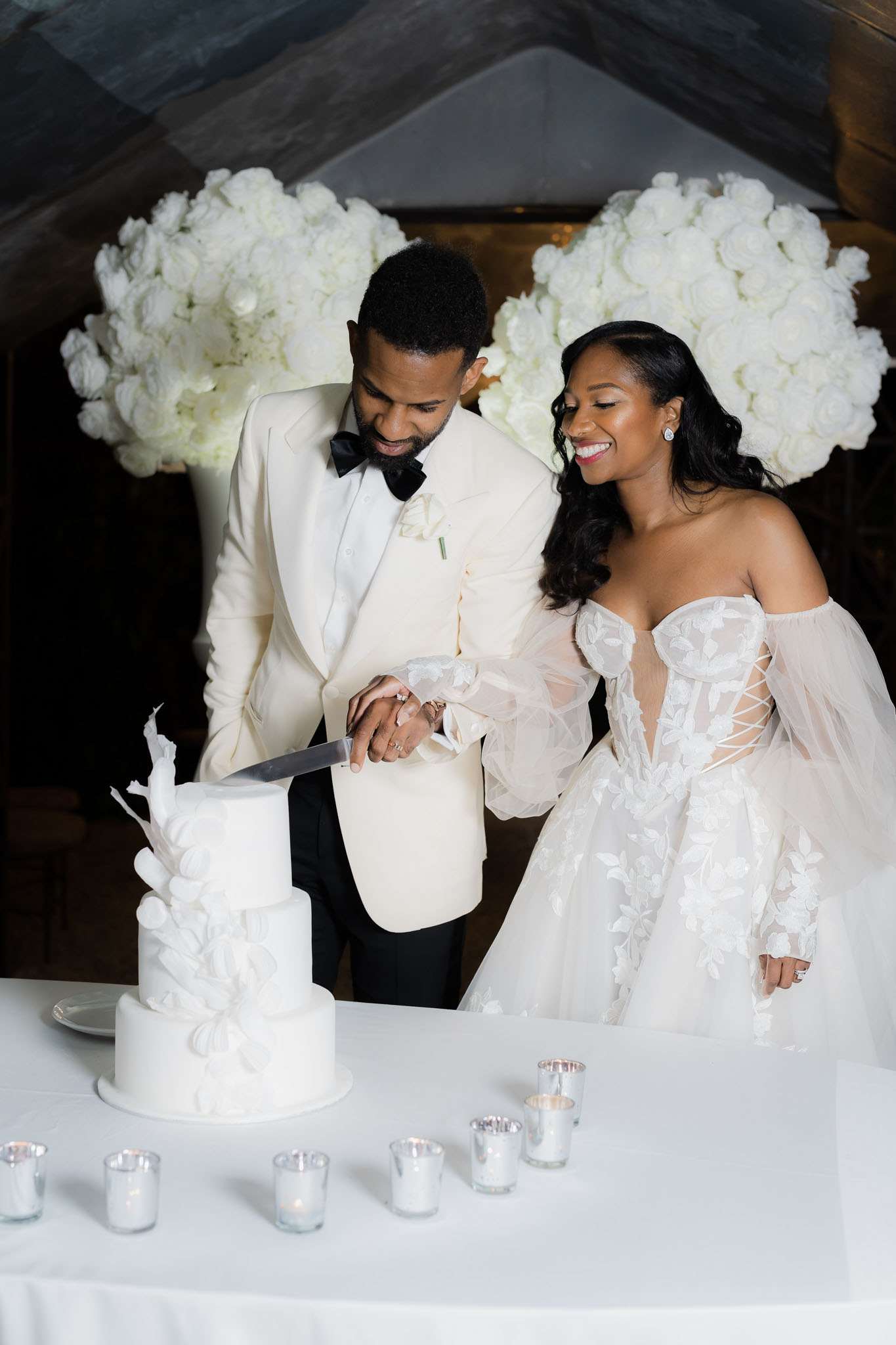 The bride and groom are cutting their wedding cake during an indoor reception, both dressed in an all-white and ivory color palette. The groom wears an ivory dinner jacket with black trousers and a black bow tie, while the bride wears an off-the-shoulder ballgown with floral lace appliqué, a corset bodice, and voluminous tulle sleeves. The three-tier all-white cake features sculptural sugar leaf and feather decorations. The table is dressed in white linen and lined with small votive candles, and two large rounded arrangements of white roses and white hydrangeas flank the couple in the background, reinforcing the monochromatic white decor theme. The shot is a mid-length portrait composition with the couple smiling and looking down at the cake.