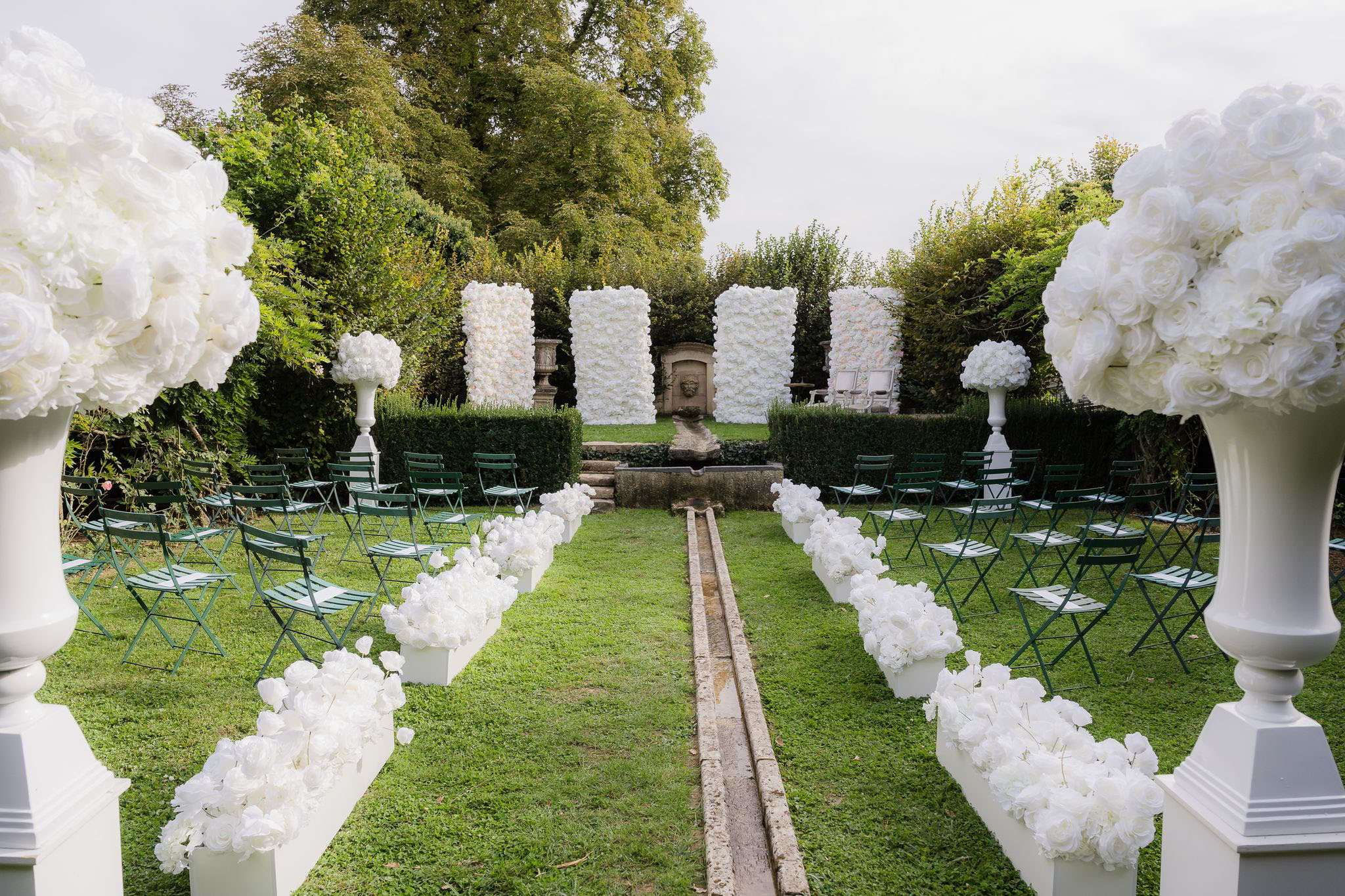 An outdoor wedding ceremony setup photographed before guests arrive, shot from a wide angle at the entrance of the aisle. The ceremony space is set within a formal garden featuring a central stone water channel running down the middle of the lawn, flanked by two rows of dark green metal folding chairs arranged in a curved layout. The aisle is lined with white floral arrangements — large bunches of white roses and hydrangeas — displayed in white rectangular planters. At the entrance, two oversized white urn pedestals hold large spherical arrangements of white roses and hydrangeas. The altar backdrop consists of three white flower walls composed of densely packed white roses, framing a classical stone fountain with a sculpted face mask, set against a neatly trimmed tall hedge. Two additional white floral topiary arrangements on white pedestal urns flank the altar area. The overall decor palette is entirely white against deep green, creating a classic formal style. Potential venue feature image.