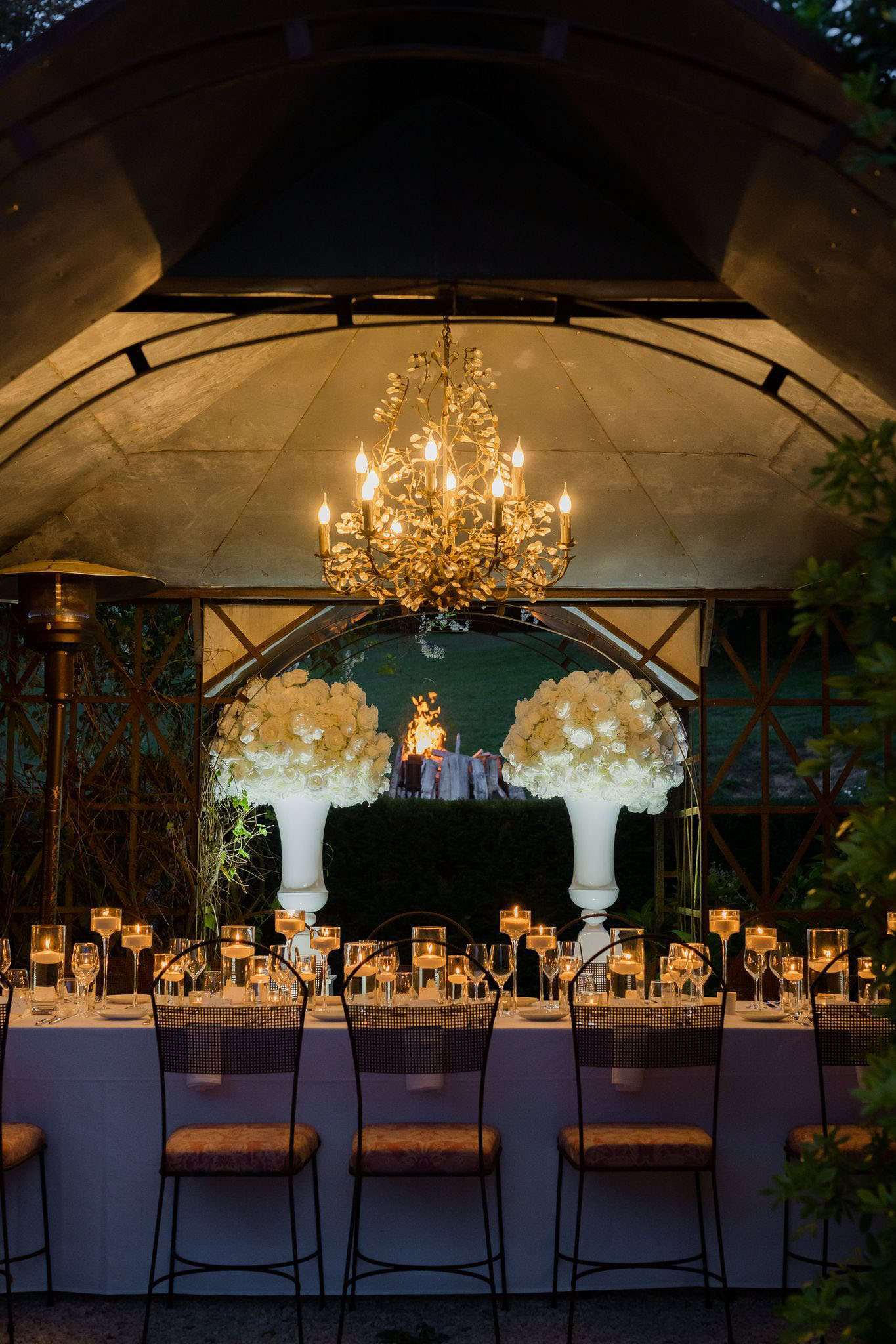 An outdoor evening reception table setup photographed at dusk under a stone-vaulted pavilion with an ornate gold leaf-motif chandelier overhead. A long rectangular dining table is dressed in a white linen tablecloth and lined with black wrought-iron cane-back chairs with tan upholstered seats. The table is set with crystal wine glasses and rows of floating candles in tall glass holders creating warm, low candlelight along the entire length. At the far end of the table, two tall white pedestal vases hold large dome-shaped arrangements of white roses and white hydrangeas. Behind the floral arrangements, a circular arch frames a view of an open-air fire pit burning in the background garden. The overall decor palette is white and gold with a classic, formal style. Wide establishing shot.