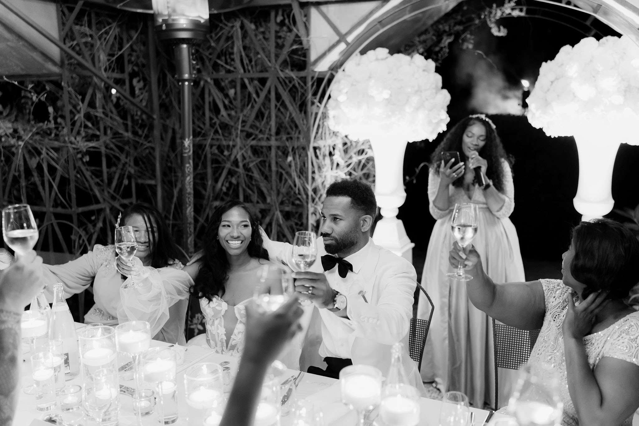 This black-and-white image captures a toast during a wedding reception, with the bride and groom seated at the head table raising champagne glasses alongside several guests. The bride wears a strapless gown with floral appliqué and sheer puff sleeves, while the groom is dressed in a white dinner jacket with a black bow tie. In the background, a woman in a light-colored long-sleeve gown stands holding a microphone and a phone, appearing to deliver a speech in front of a large white floral installation composed of oversized blooms arranged in a circular arch. The table in the foreground is set with floating candle centerpieces and glassware, and the venue features a decorative trellis backdrop with trailing branches, suggesting an outdoor or covered terrace setting. The image is a medium wide shot with high contrast and bright highlights on the glassware and floral backdrop.
