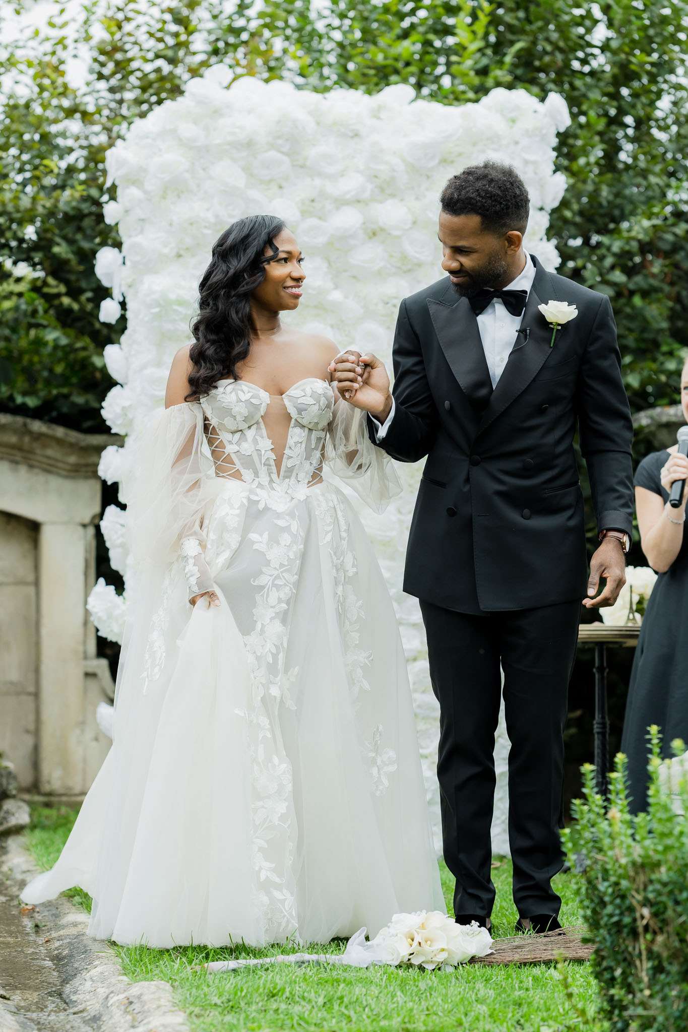 A couple stands together at the altar during an outdoor wedding ceremony, sharing a joyful moment as the groom holds the bride's hand and looks down at her. The bride wears an ivory tulle ball gown with a deep sweetheart neckline, 3D floral appliqué leaf detailing along the bodice and skirt, a lace-up corset front, and sheer off-the-shoulder puff sleeves; a loose bouquet of white blooms rests on the ground at her feet. The groom wears a black double-breasted tuxedo with a bow tie and a white rose boutonniere. Behind them is a large rectangular white rose wall backdrop, and a partially visible officiant stands to the right. The setting appears to be a garden or chateau grounds, and the overall decor palette is all-white with a modern classic style. Medium two-shot portrait composition.