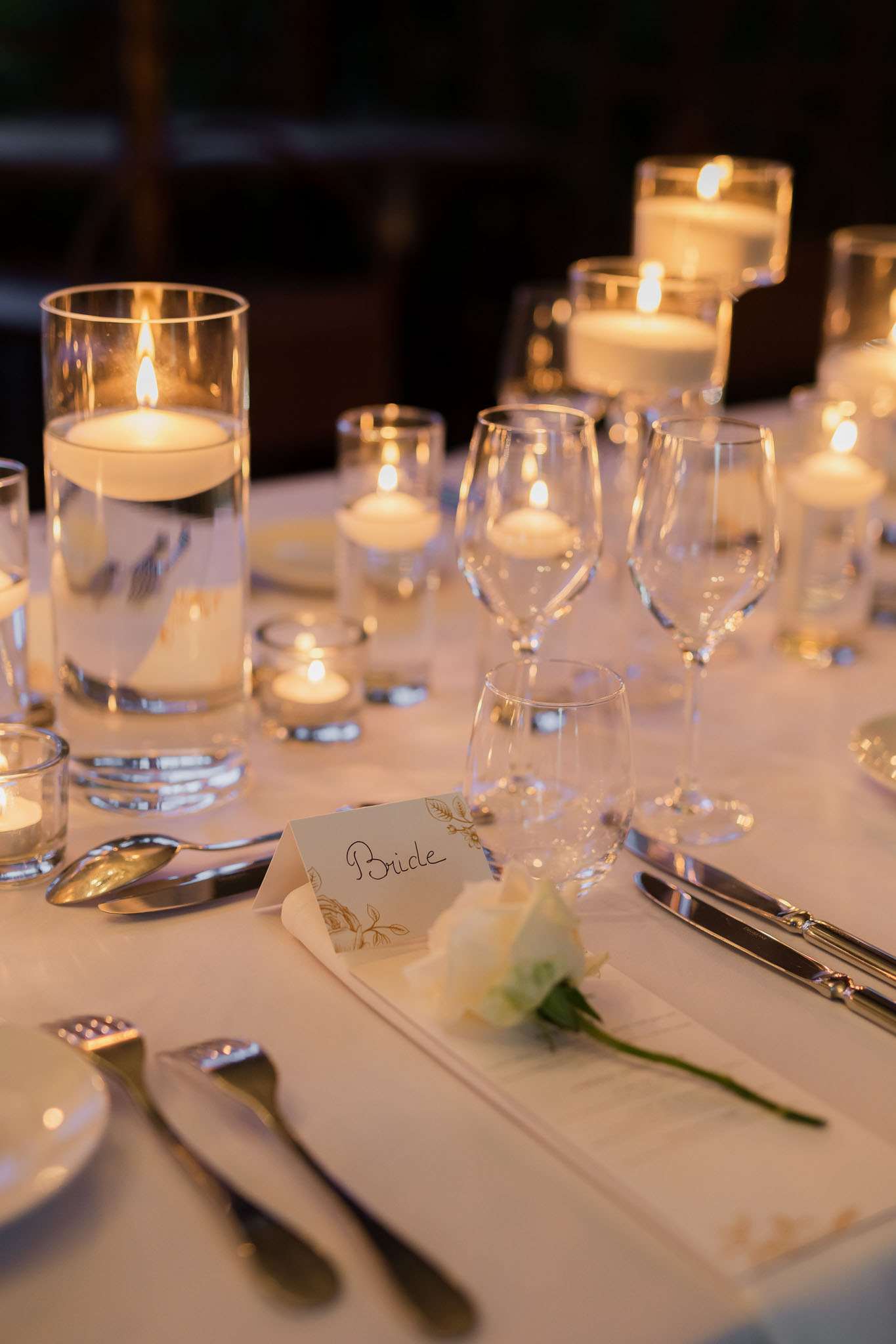 Close-up detail shot of a wedding reception table setting, photographed indoors in a dimly lit room. The table is covered with a white linen cloth and set with silver cutlery, clear wine glasses, and white folded napkins. A handwritten place card reading 'Bride' with gold botanical illustration detailing is positioned on the napkin alongside a single ivory rose stem. The centerpiece consists of multiple floating candles in tall and low cylindrical glass vessels, along with small votive tealight holders scattered across the table, creating warm ambient candlelight. The overall decor palette is white, ivory, and gold with a classic, candle-lit reception style.