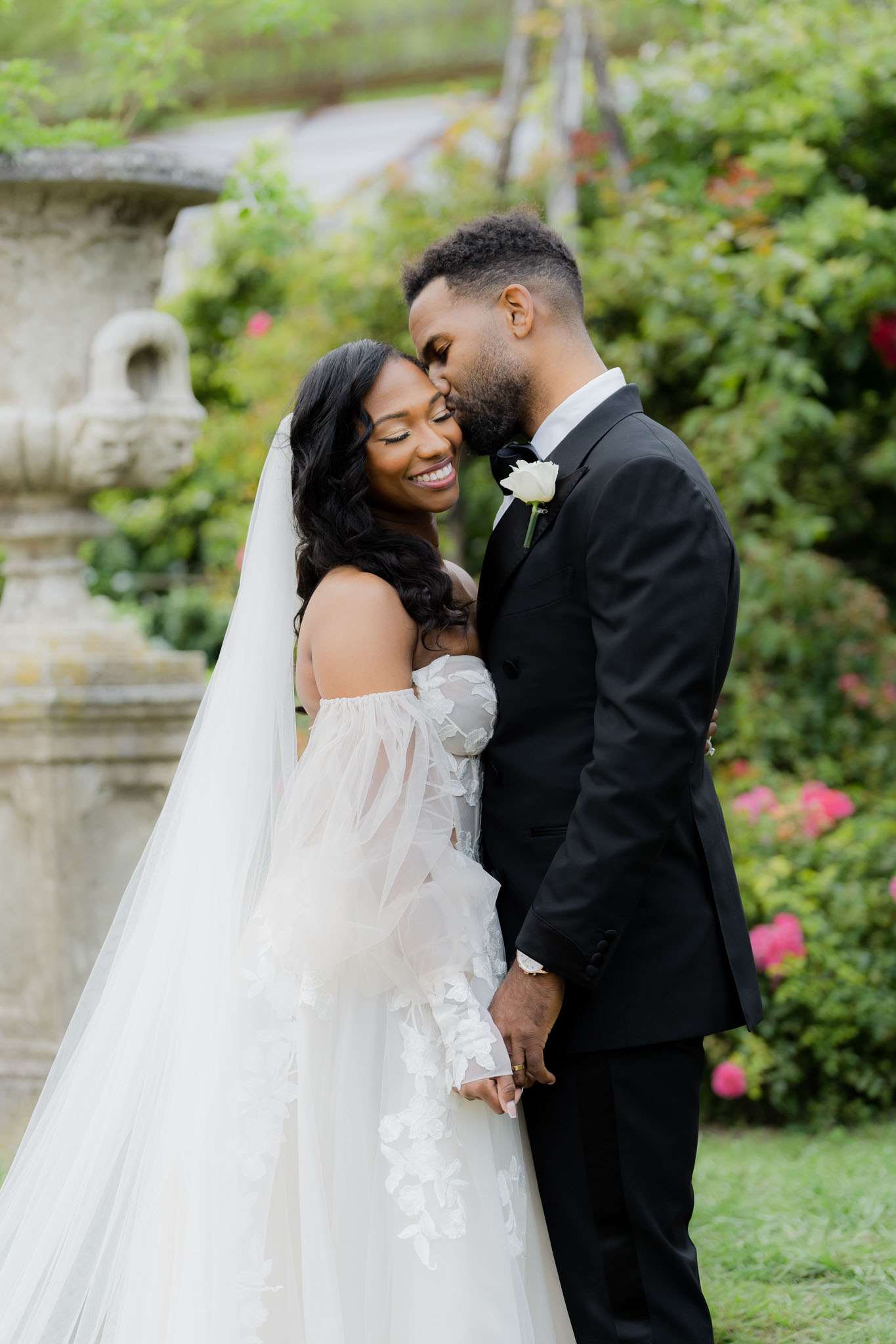 A couple portrait taken outdoors in a garden setting, with a stone urn and lush greenery visible in the background. The bride wears an off-the-shoulder white ballgown with sheer, floral appliqué long sleeves, a cathedral-length veil, and her dark hair in loose waves; she is smiling broadly with her eyes closed as the groom nuzzles her cheek. The groom is dressed in a black tuxedo with a black bow tie and a single white rose boutonniere. The composition is a medium portrait shot with the couple centered and the background softly blurred, conveying a classic black-and-white color palette in the styling choices against the garden backdrop.