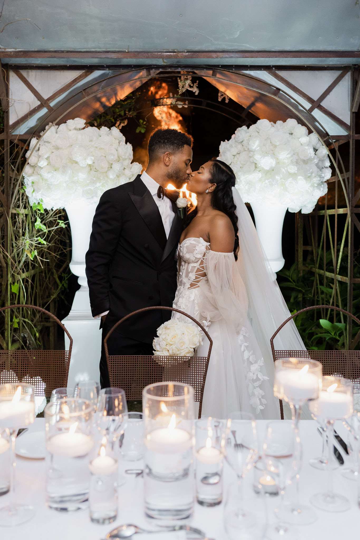 A bride and groom share a kiss at an outdoor evening reception, framed by a circular iron arch flanked by two large white urn arrangements densely filled with white roses. The groom wears a black tuxedo with a white boutonniere, while the bride wears a strapless corset-style gown with floral appliqué detailing, a lace-up front, off-the-shoulder tulle sleeves, and a long veil; she holds a compact round bouquet of white roses. The foreground features a white-linen reception table set with numerous pillar and votive candles in glass vessels, wine glasses, and silverware, all contributing to a candlelit, all-white and black color palette with a classic, formal styling theme. This is a medium portrait shot taken from across the reception table, with the warm backlit glow from behind the arch creating depth in the background.