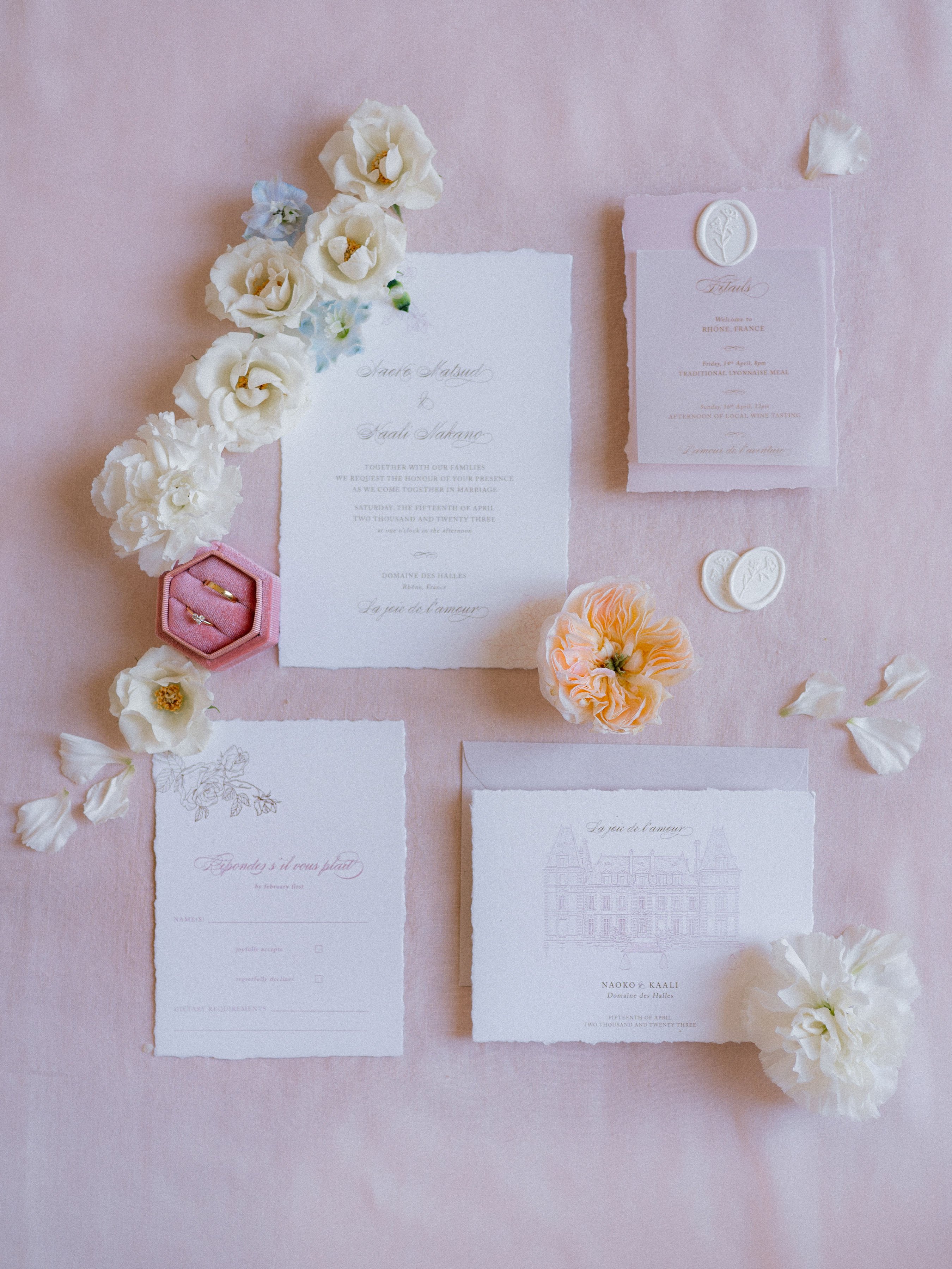 A flat lay detail shot of a complete wedding stationery suite arranged on a blush pink linen background. The suite includes a main invitation with deckled edges printed in soft grey calligraphy script, a details card and RSVP card both on matching deckled-edge paper, a venue illustration card featuring a line drawing of Domaine des Halles in the Rhône, France, a grey envelope, and two ivory wax seals with floral motifs. Scattered around the stationery are cream lisianthus blooms, a pale peach garden rose, soft blue delphinium sprigs, and loose ivory petals. A dusty pink hexagonal velvet ring box holding two gold wedding bands is positioned to the left of the main invitation, adding a jewelry detail to the composition. The overall palette is blush, ivory, and soft grey with a classic, French-inspired romantic styling.