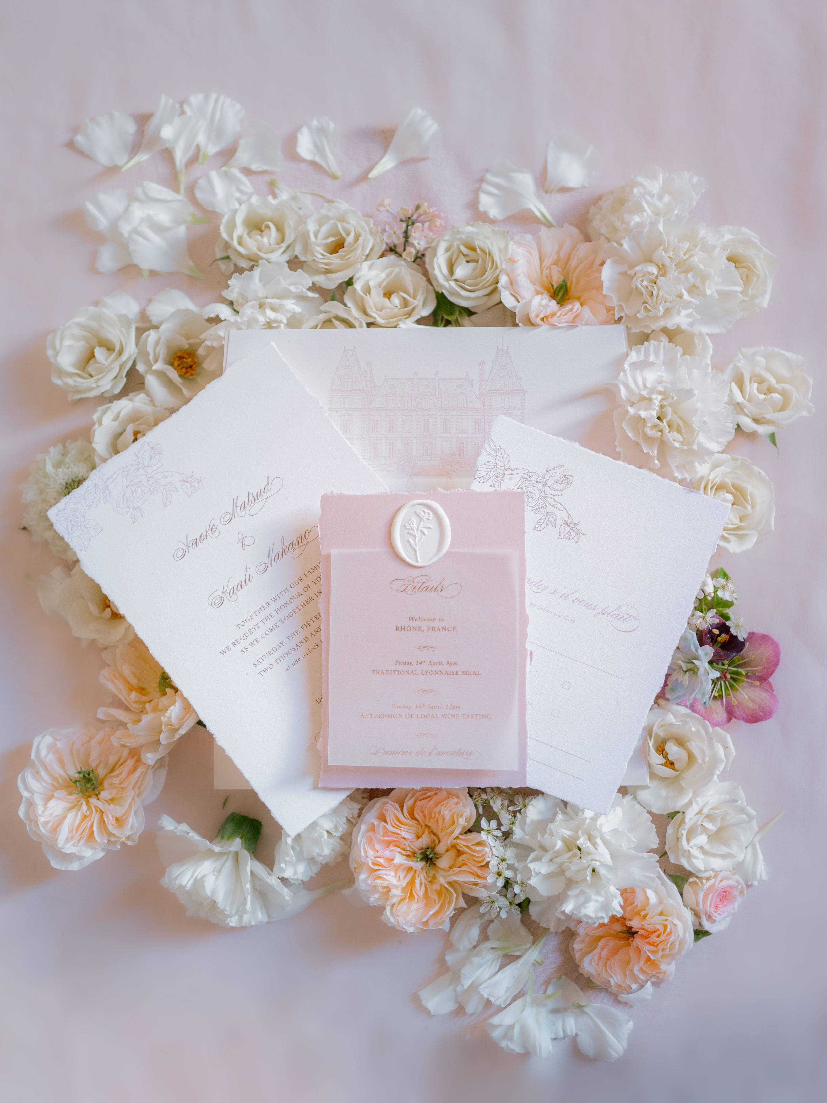 A flat lay detail shot of a wedding stationery suite arranged on a blush pink fabric background, surrounded by loosely scattered ivory garden roses, peach David Austin roses, white carnations, and small blush pink blooms with scattered petals. The suite includes a white deckle-edge invitation with calligraphy script and floral illustration, a white envelope featuring a line-drawing illustration of a French chateau or manor house, a blush pink details card sealed with a cream floral wax seal, and a white RSVP card. The details card references a wedding in Rhone, France, with events including a Traditional Lyonnaise Meal and an Afternoon of Local Wine Tasting. The overall palette is blush pink, ivory, and warm gold, with a classic and romantic stationery design.