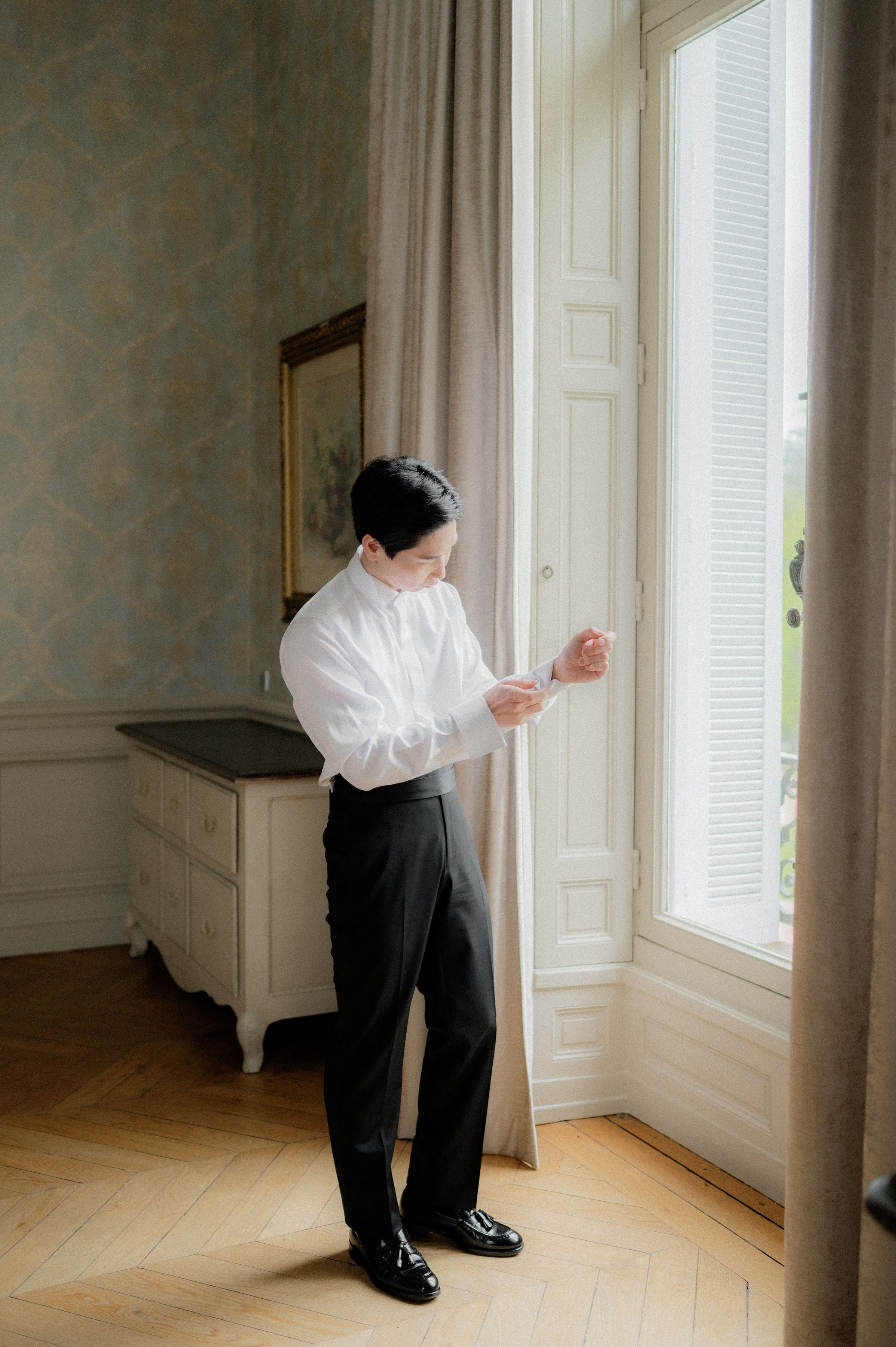 A groom getting ready in an ornate chateau interior, fastening his cufflinks near a tall French door with louvered shutters. He is dressed in a white tuxedo shirt with a bib front, black high-waisted trousers, a black cummerbund, and black tassel loafers. The room features blue-green damask wallpaper, a cream-painted chest of drawers with a dark marble top, a gold-framed painting, herringbone parquet flooring, and draped taupe curtains. The shot is a full-length portrait framed by natural light streaming through the open door.
