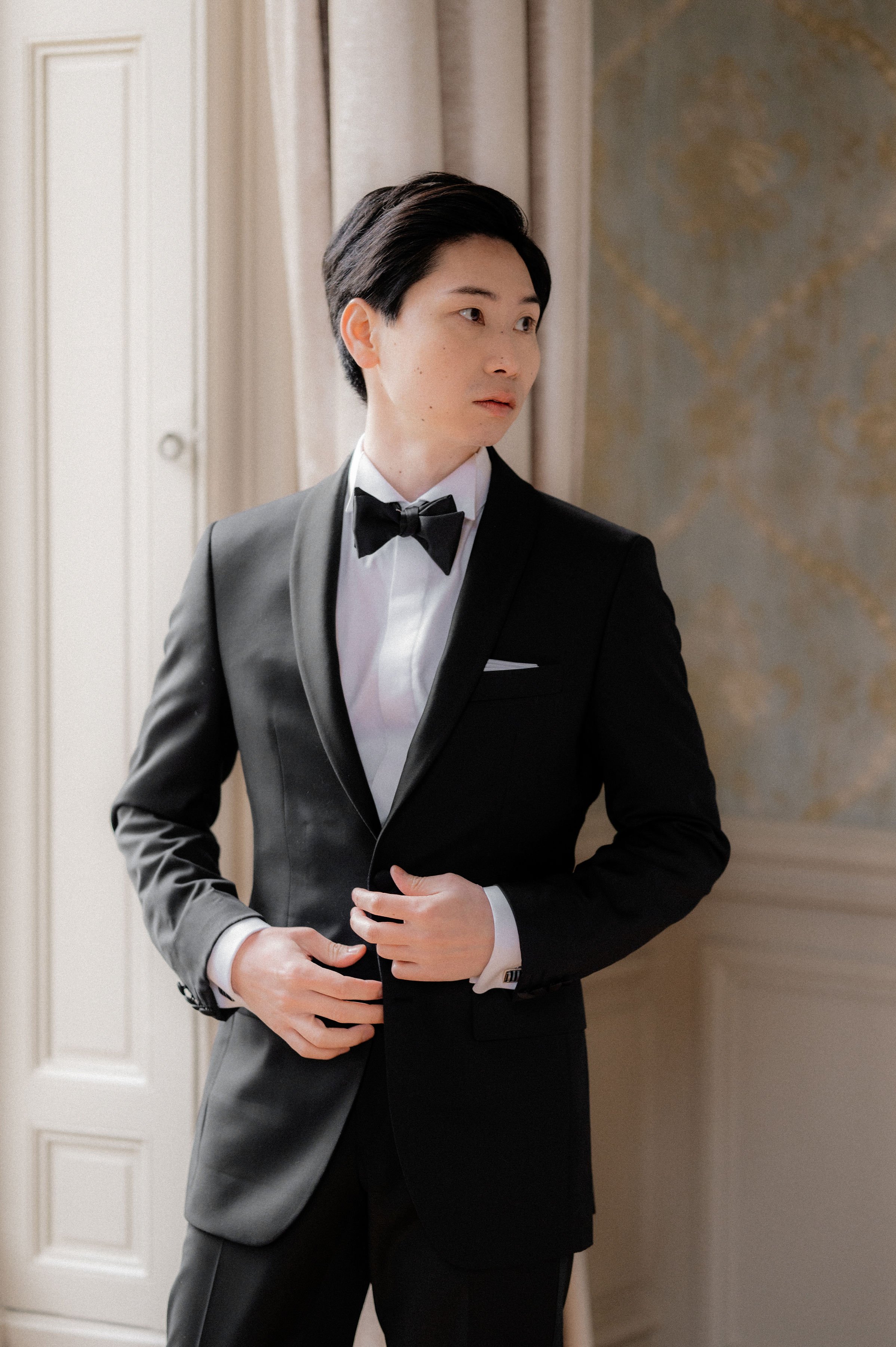 A getting-ready portrait of a groom standing indoors in a formal room with ornate gold-and-grey wallpaper and a white paneled door with floor-length cream curtains. He is wearing a black tuxedo with a satin shawl lapel, white dress shirt, black bow tie, white pocket square, and cufflinks, and is adjusting the front of his jacket while gazing to one side. The shot is a close-up portrait framed from roughly mid-thigh up, with soft natural light coming from the left side of the frame, giving a classic formal aesthetic.