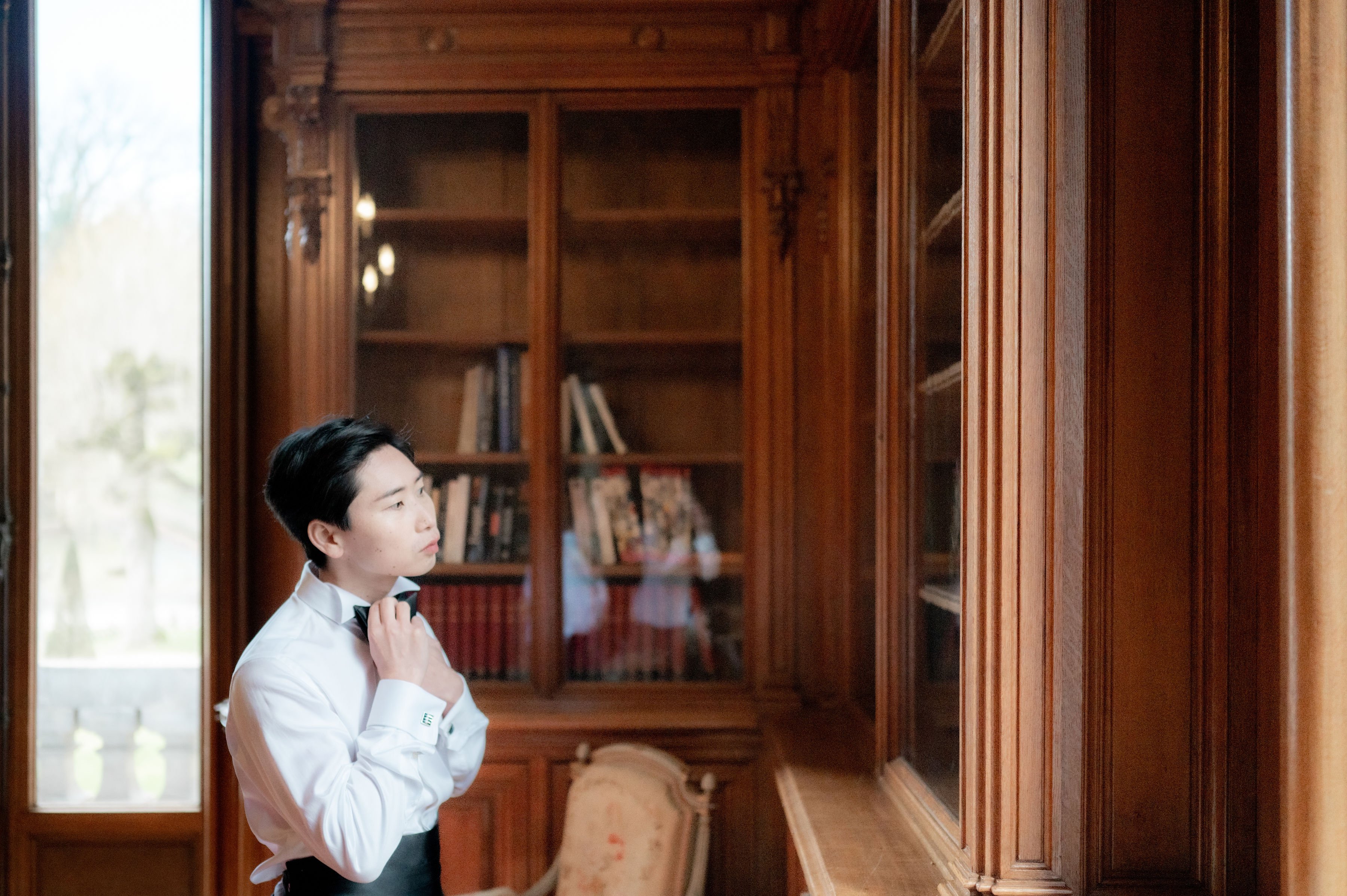 A groom getting ready indoors, adjusting his black tie while glancing to the side toward a tall window. He is wearing a white dress shirt with French cuffs and dark trousers. The setting is a richly paneled wooden library room, likely within a chateau, featuring floor-to-ceiling carved walnut bookshelves filled with books, glass-fronted cabinet doors, ornate column detailing, and a small antique upholstered chair visible in the background. Warm pendant lights glow softly inside the bookcase. The portrait is a medium shot with natural window light illuminating the subject from the left, creating a classic getting-ready moment within a formal, traditional interior.