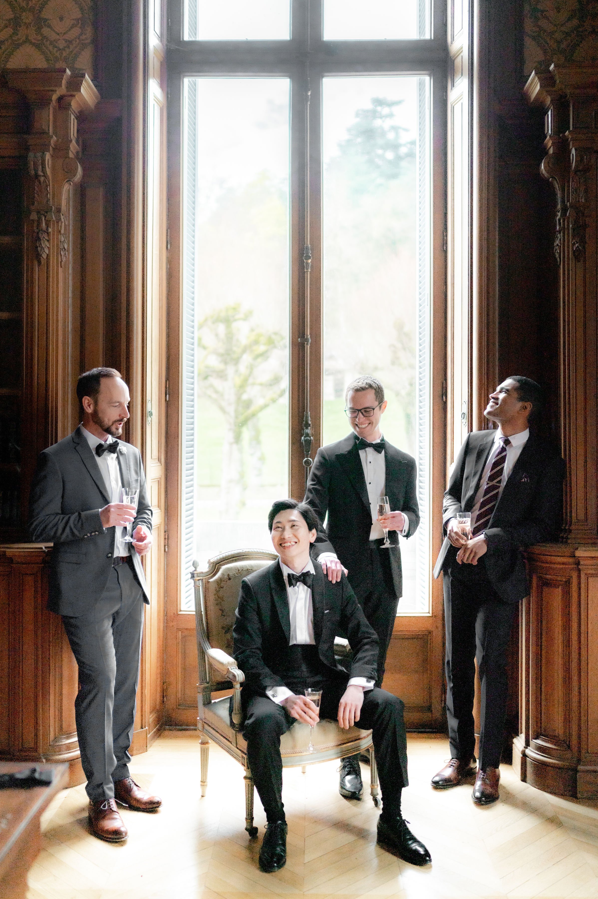 Four men are gathered indoors in a getting-ready or pre-ceremony moment, holding champagne flutes and laughing together in a relaxed group portrait. The setting is an ornate interior room of what appears to be a French chateau, featuring tall carved dark wood paneling, decorative pilasters, and a large floor-to-ceiling French window that floods the scene with natural backlight. The groom, seated on a gold Louis XVI-style armchair, wears a black tuxedo with a bow tie; two other men also wear black tuxedos with bow ties, while the fourth wears a charcoal grey suit with a striped tie. The composition is a full-length portrait shot with the window centered behind the group, creating a bright contrast against the warm wood tones of the room.